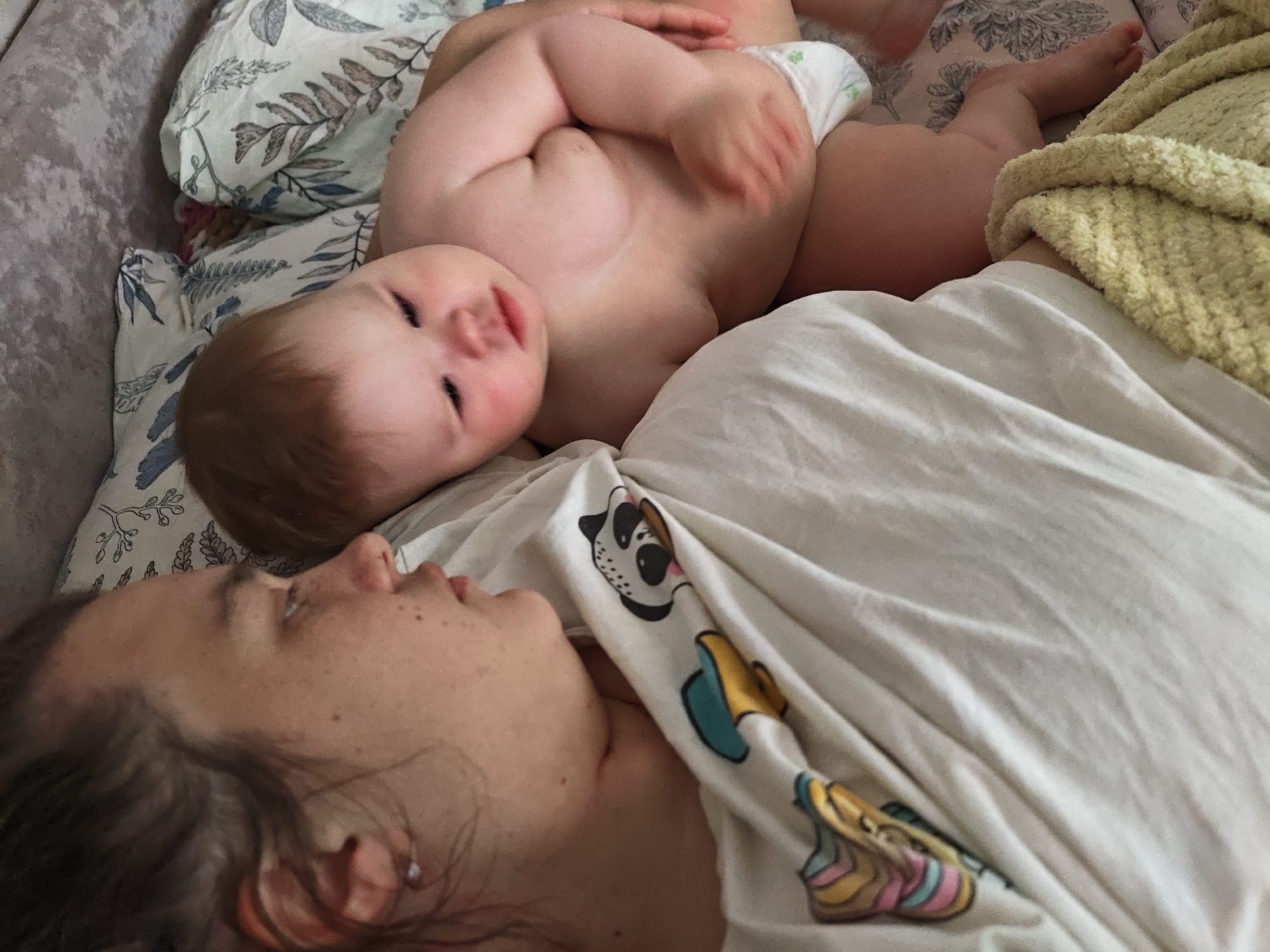 The image shows a woman and a baby lying on a bed or sofa. The woman is lying on her back, facing away from the camera. The baby is lying on top of the woman, facing the camera. The baby is wearing a diaper. The woman is wearing a white t-shirt with cartoon images. The bed or sofa has a floral-patterned pillow. A yellow blanket is visible next to the woman. The location is unspecified, despite the query suggesting Chaikovsky, Russia, as there are no indicators in the visual data to verify this.
