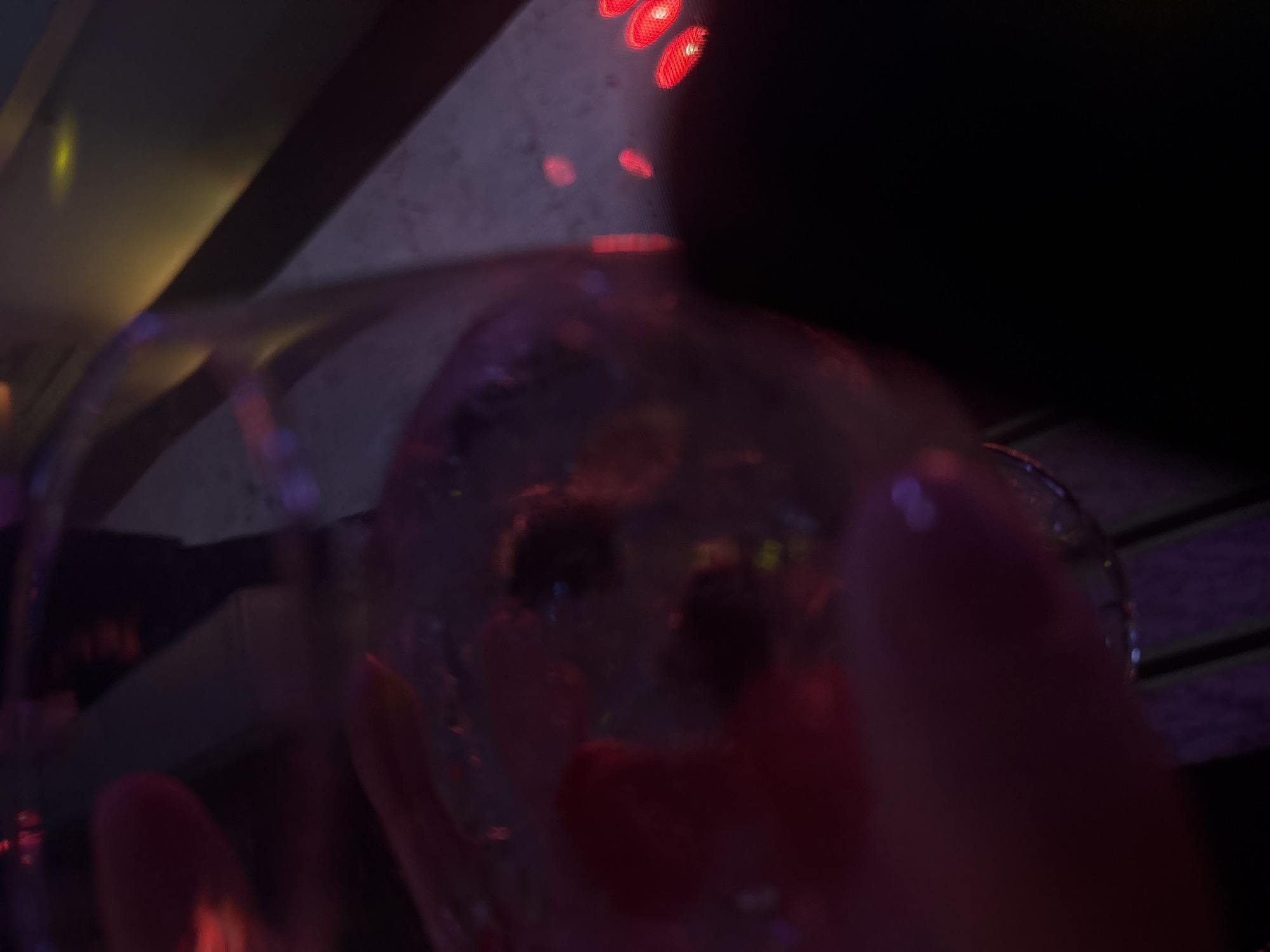 The image presents a highly blurred and underexposed scene, predominantly colored by red and pink ambient light. A clear stemmed glass, containing a translucent liquid with suspended dark and red fruit pieces (likely berries) and small reflective particles, is prominently featured in the central-left, held from below by an indistinct light-skinned hand. The background is dark and amorphous. Visible elements include several distinct red, oval-shaped lights arranged horizontally on the right side, along with diffuse purple and yellow light reflections. Vertical linear elements, consistent with structural supports or railings, are vaguely discernible in the background. The setting is an indoor, low-light environment, consistent with a bar, restaurant, or lounge. No specific geographical markers or landmarks are present to identify a precise location within Mexico City, Mexico.