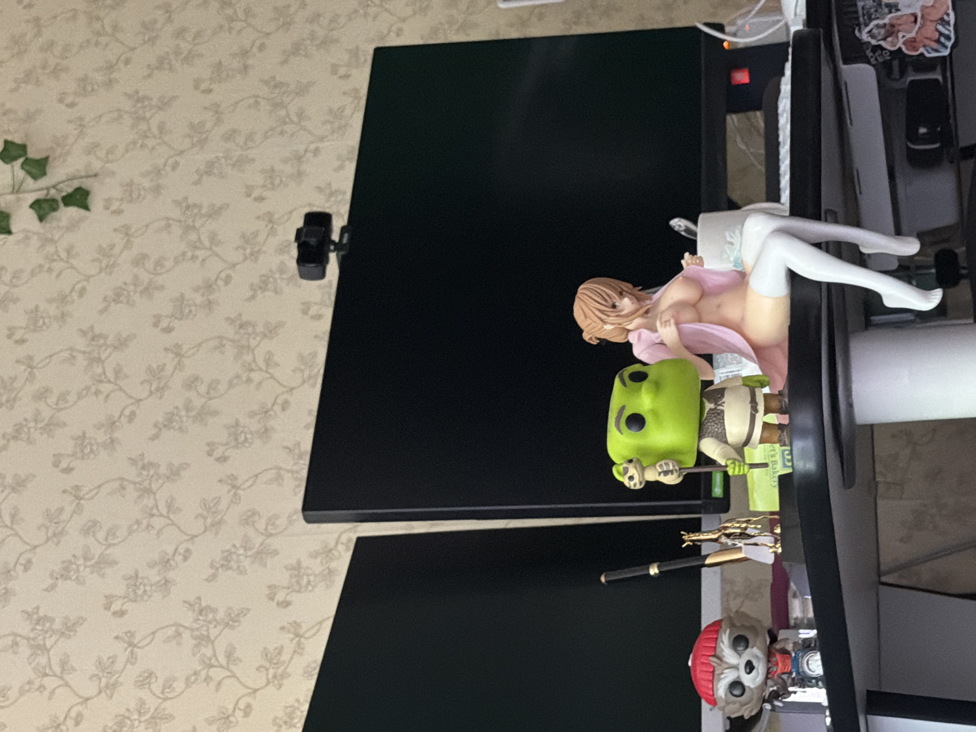 This is a photograph of a desk with two computer monitors. On the desk are several figurines, including one of Shrek, one of Rocket Raccoon from Guardians of the Galaxy, and an anime-style figurine of a woman. There is also a webcam on top of one of the monitors. The background is wallpaper with a floral pattern. The setting is a room in Khimki, Russia.