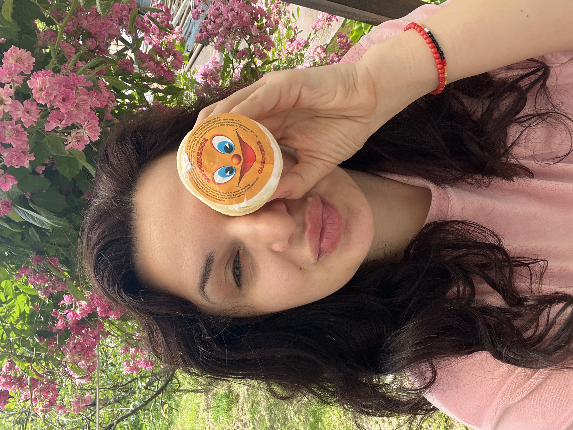 A woman with long, dark, wavy hair is depicted outdoors, holding a circular object to her right eye. She is wearing a light pink garment and has her left eye partially closed in a wink, while her lips are puckered. Her left wrist displays two beaded bracelets, one predominantly red, and the other multi-colored with red and black beads. The object held is an orange-colored wrapper for what appears to be ice cream, featuring a smiling cartoon face with blue eyes and a red nose, and Cyrillic text, including "МОРОЖЕНОЕ" (Ice Cream) and "САМОВАРЧИК" (Samovarchik). The background is filled with vibrant pink blossoms from a flowering shrub or tree, interspersed with green foliage. To the far left, a grassy area and additional greenery are visible, while to the far right, a light-colored structure can be faintly seen behind the flowers. The scene is illuminated by natural light in an outdoor setting in the city of Unknown, Unknown.