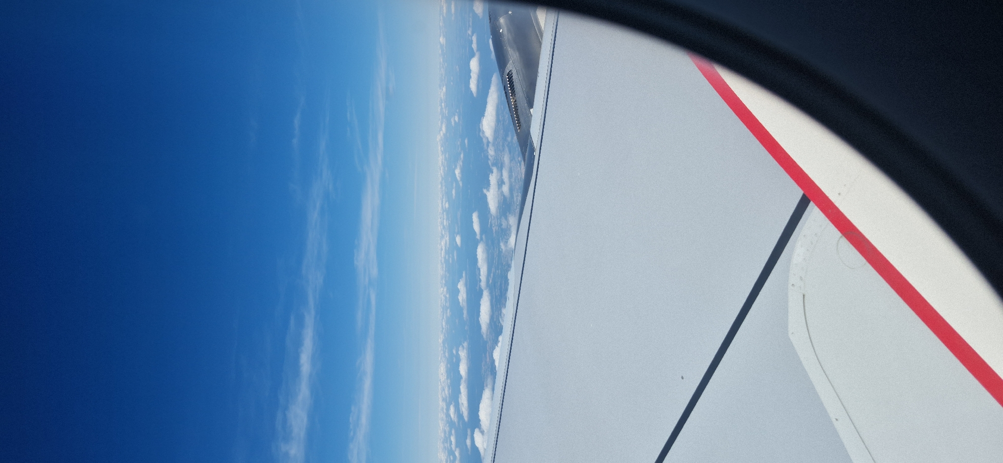 The image is an aerial view from an airplane window, looking out at the sky and clouds. The airplane's wing is prominently visible in the foreground, with a white surface and a red stripe along its edge. The sky is a deep blue, with scattered white clouds forming a layer above the horizon. The environment appears to be clear and sunny, with good visibility. There are no people or specific landmarks identifiable that would indicate the location in Groß-Zimmern, Germany. The image captures a serene moment during an airplane journey.