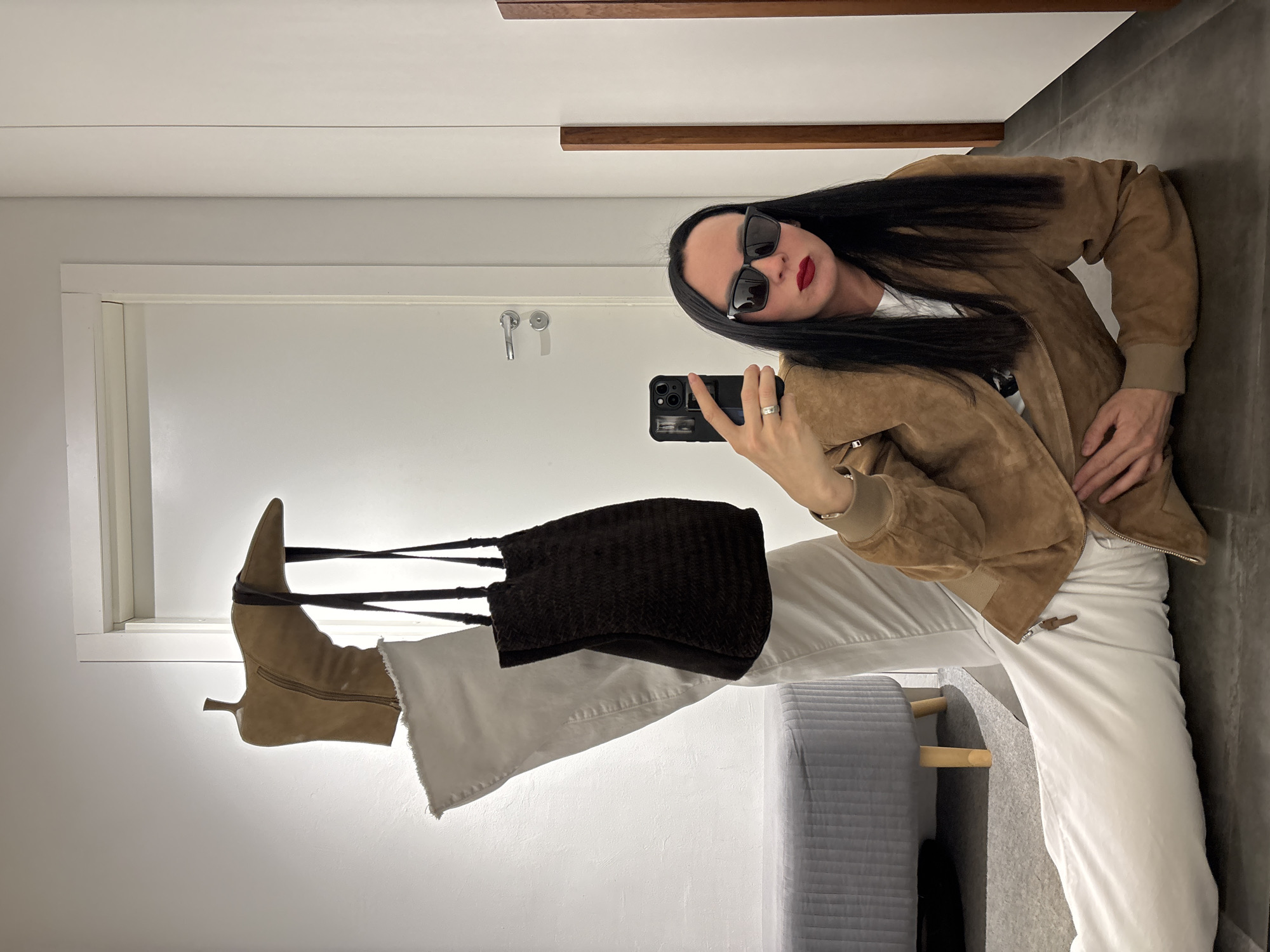 A woman with long black hair, wearing dark sunglasses, a brown suede jacket, and white pants, is taking a selfie. She is holding a black smartphone in her right hand, which is adorned with a silver ring. In her left hand, she is holding a brown corduroy tote bag, which dangles alongside a light brown ankle boot. The background suggests an interior space with a white door and window, a grey stool, and a tiled floor. There is no indication that the scene is in Chaikovsky, Russia.