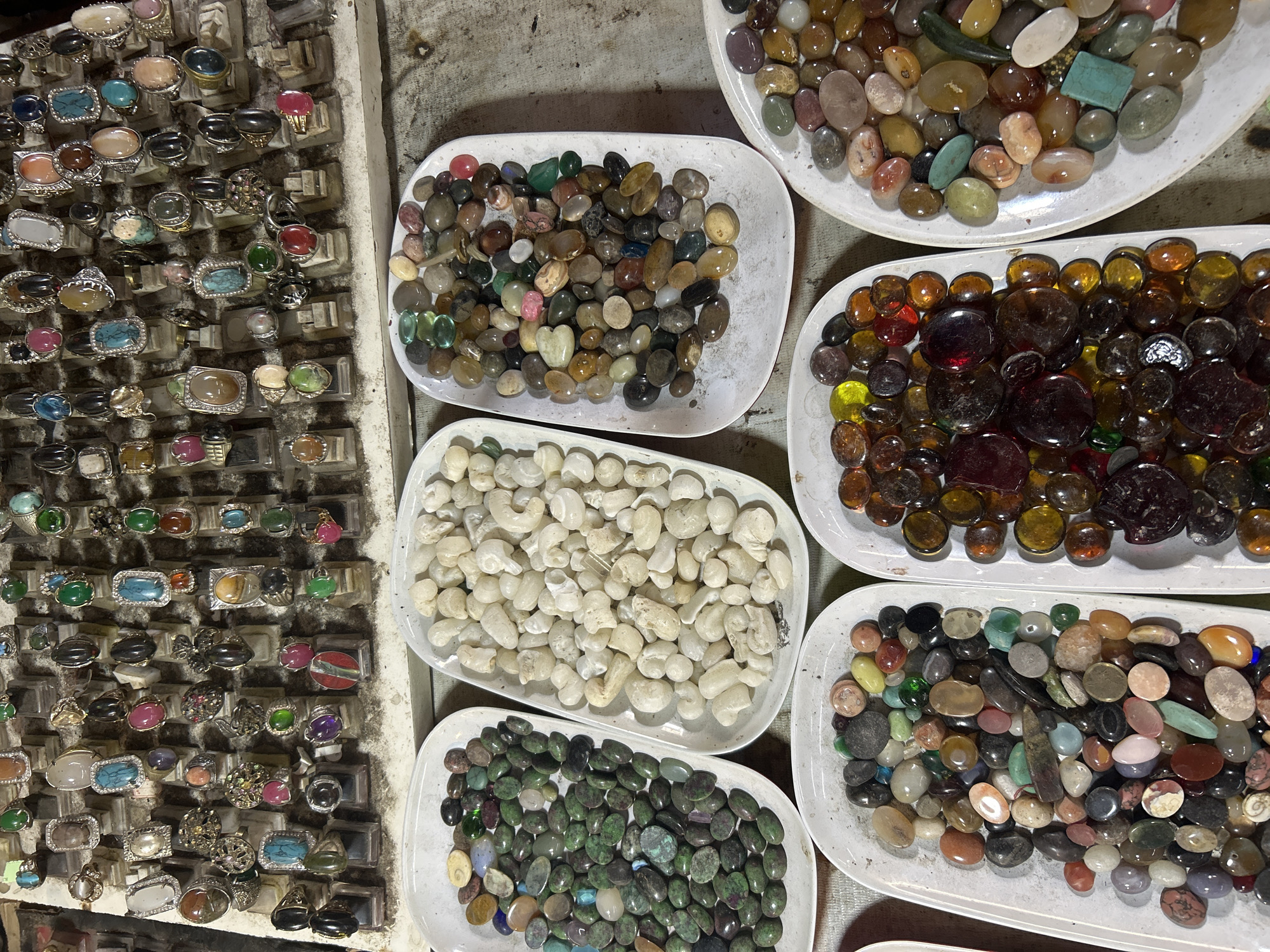 The image displays a close-up, overhead view of a market stall in Denpasar, Indonesia, featuring a large assortment of jewelry and decorative stones. In the background, rows of rings with various gemstones are neatly arranged on display stands. The rings exhibit diverse stone colors and settings, including turquoise, red, green, and clear stones, along with intricate metalwork.
In the foreground and middle ground, several white trays are filled with numerous tumbled stones. These stones vary in size, shape, color, and translucency. Some trays contain predominantly green and dark green stones, interspersed with reddish and lighter-colored stones. Another tray is filled with pale, creamy white stones that appear to be carved or shaped. Other trays showcase a mix of earthy tones, including browns, grays, oranges, and yellows, alongside translucent red, purple, and yellow stones. A tray on the far right contains a collection of predominantly brown and orange rounded glass-like objects, possibly marbles or decorative cabochons.
The overall scene suggests a vendor selling a wide variety of jewelry and decorative stones at a market. There are no people visible in the frame, and the focus is entirely on the merchandise. The lighting is consistent with an indoor or shaded outdoor market environment.