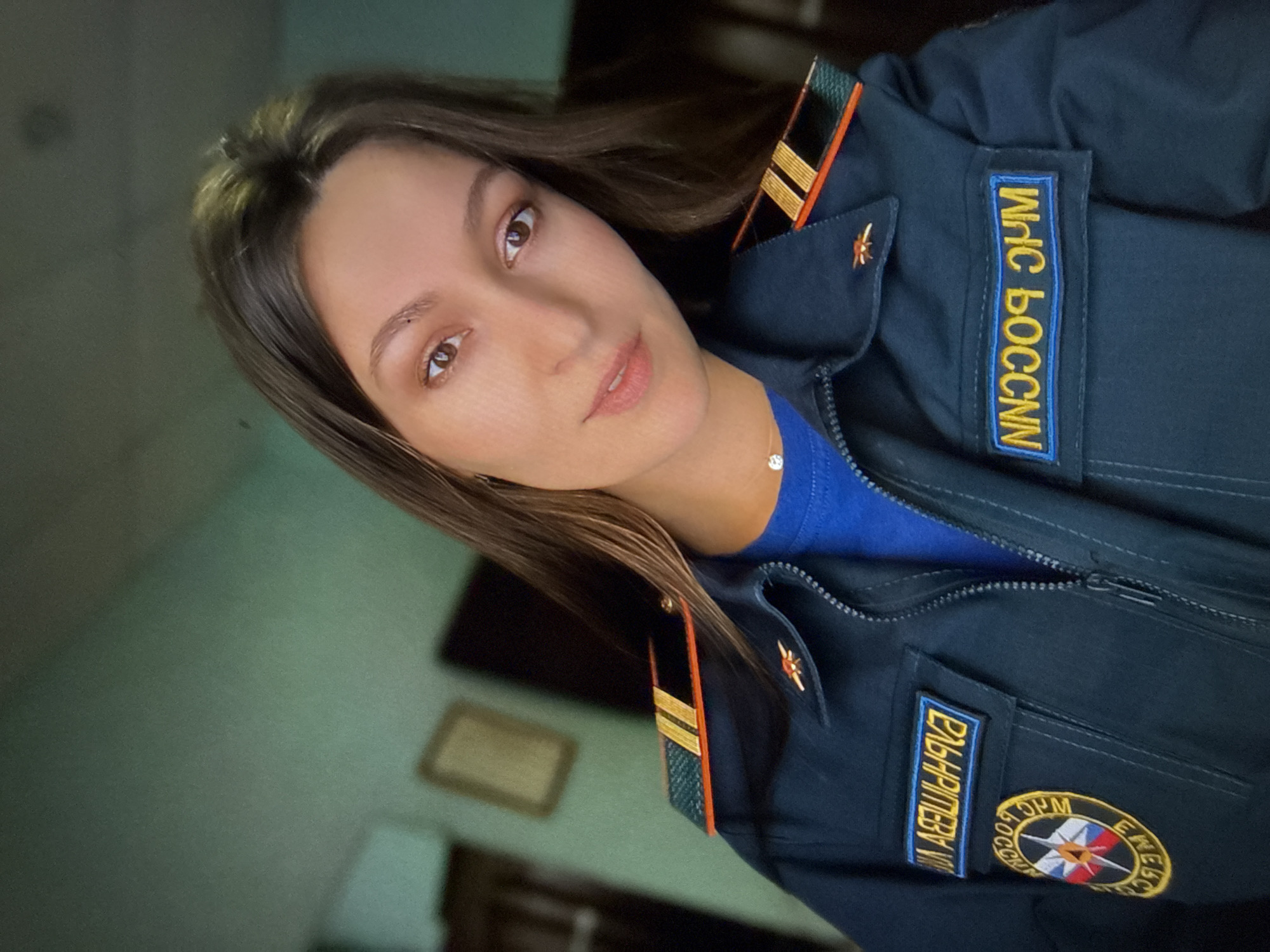The image is a close-up, selfie-style portrait of a young woman wearing a dark blue uniform jacket with gold and orange epaulets. She has long, dark brown hair and is looking directly at the camera with a neutral expression. The jacket has two patches: one on the left chest with a circular emblem and text, and another on the right chest with yellow text on a blue background. The background is blurry and appears to be an indoor setting with muted green walls and a framed picture on the wall. There is no discernible information in the image that would place the location in the city of Chaikovsky, Russia.