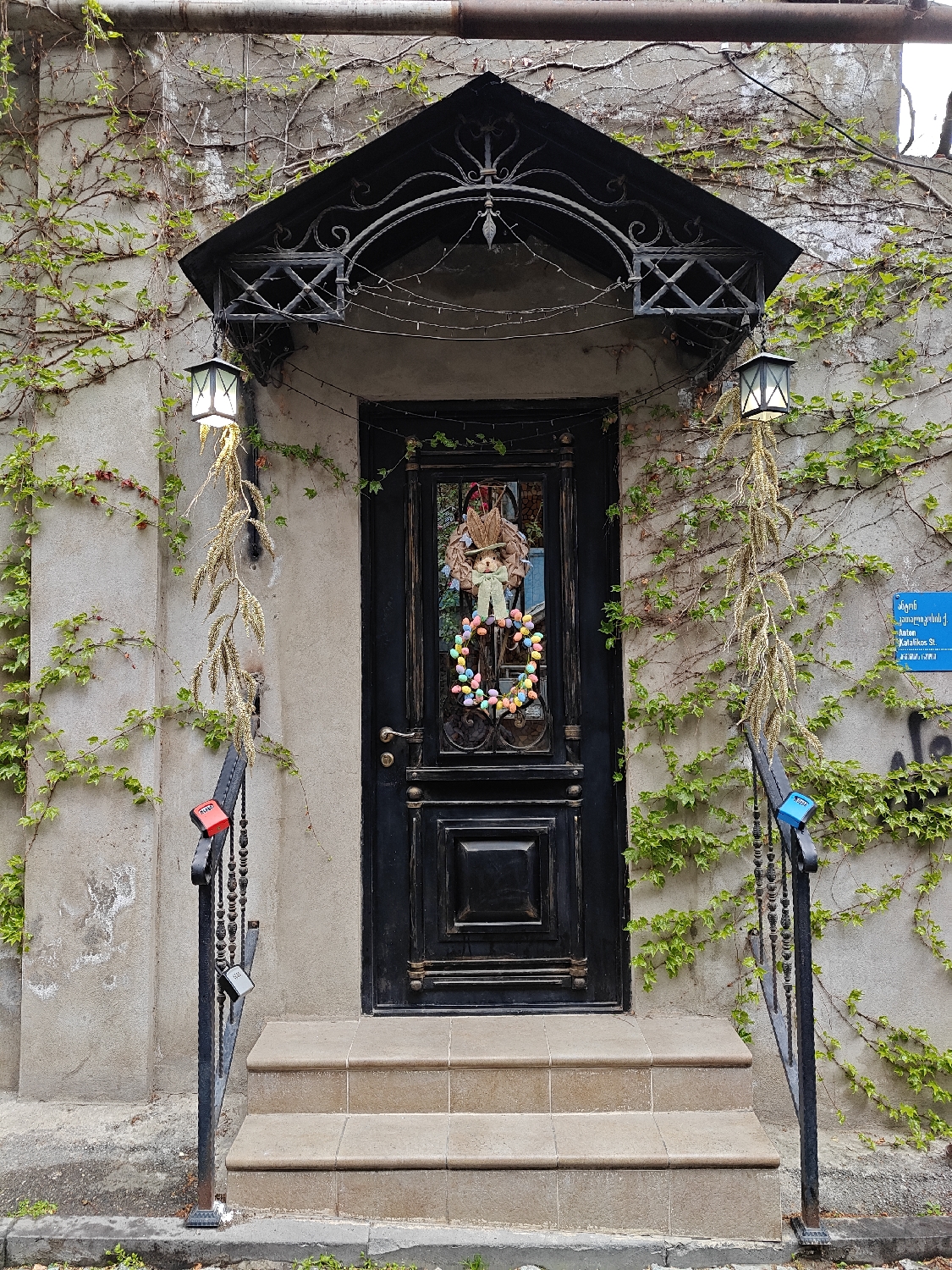 The image captures a charming and decorated doorway of a building, likely in Tbilisi, Georgia. The focal point is a dark, possibly black, wooden door with raised panels, set into a light-colored concrete or stucco facade. An ornate, black wrought-iron canopy arches over the door, from which two outdoor lanterns with glowing panes hang. Delicate string lights are also draped beneath the canopy.
A festive Easter-themed wreath adorns the door, featuring a plush bunny figure and a colorful ring of pastel-colored eggs. Long, dried, wheat-like stalks are gracefully draped from each lantern, enhancing the decorative appeal. The facade itself is extensively covered in green ivy and other climbing vines, giving the building a natural, weathered look.
Three worn, light brown tiled steps lead up to the door, flanked by black wrought-iron railings. Notably, several love locks are attached to the railings; a bright red one and a blue one are prominent, along with a darker one visible on the left railing.
On the right wall, a blue rectangular sign with white Georgian script is visible. The text reads: "გუმბათის / კათოლიკოსი. ქ.", which translates to "Gumbatis / Katolikos St." The scene appears to be set during the daytime under an overcast sky, suggesting soft lighting and mild weather. No people are visible.