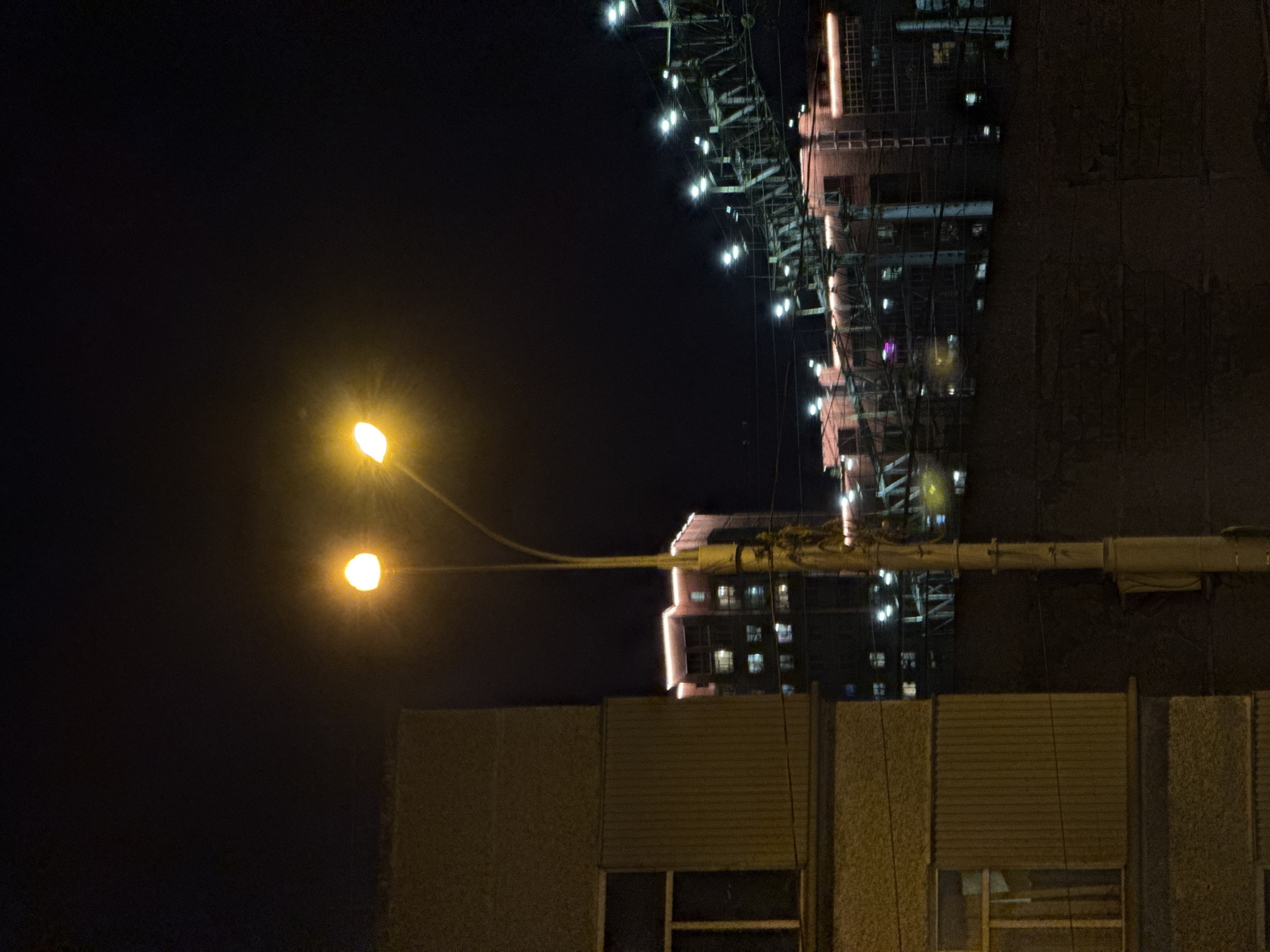 The image shows a nighttime scene in what appears to be Perm, Russia. A street lamp with two illuminated bulbs is positioned in the foreground. Behind the lamp, a building with some visible windows is partially obscured. In the background, there's a taller building with a lighting structure on top, typical of urban architecture. The overall lighting is dim, with the street lamp and building lights providing the main illumination sources. The image is rotated 90 degrees counter-clockwise.