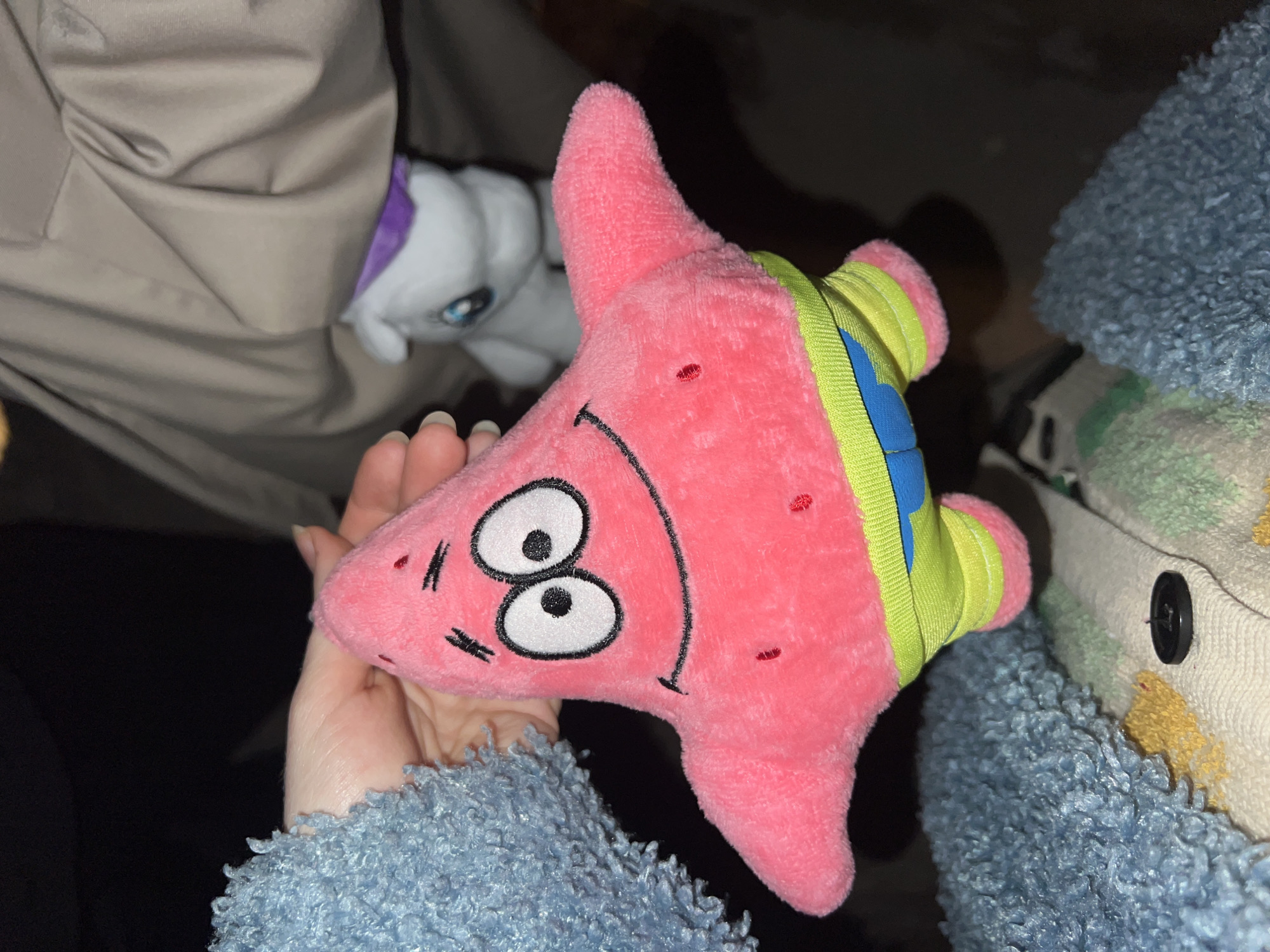 The image depicts a hand holding a pink Patrick Star plush toy. The toy has a smiling face and is wearing a green and blue accessory. To the left, there's a partial view of a light-colored garment. In the background, there's a white plush toy with purple accents and to the right is a blue, fluffy texture with a garment containing a button. The location in the city of Belgrade, Serbia, is not discernible from the image.