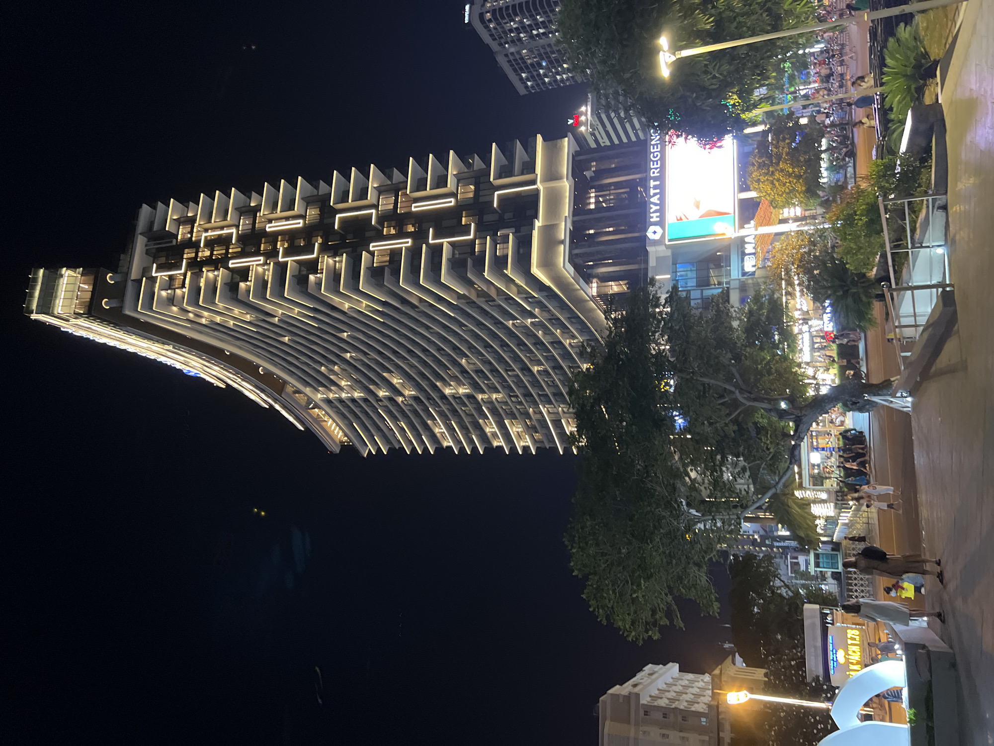 The image shows a night scene in Nha Trang, Vietnam, featuring the Hyatt Regency hotel. A large, modern, curved skyscraper with illuminated balconies dominates the center of the frame. Below the hotel, a brightly lit digital billboard displays an advertisement. In the foreground, a paved walkway is visible, with several people walking and standing. Trees and streetlights are also present, contributing to the urban nighttime atmosphere. The sky is dark, indicating it is nighttime.