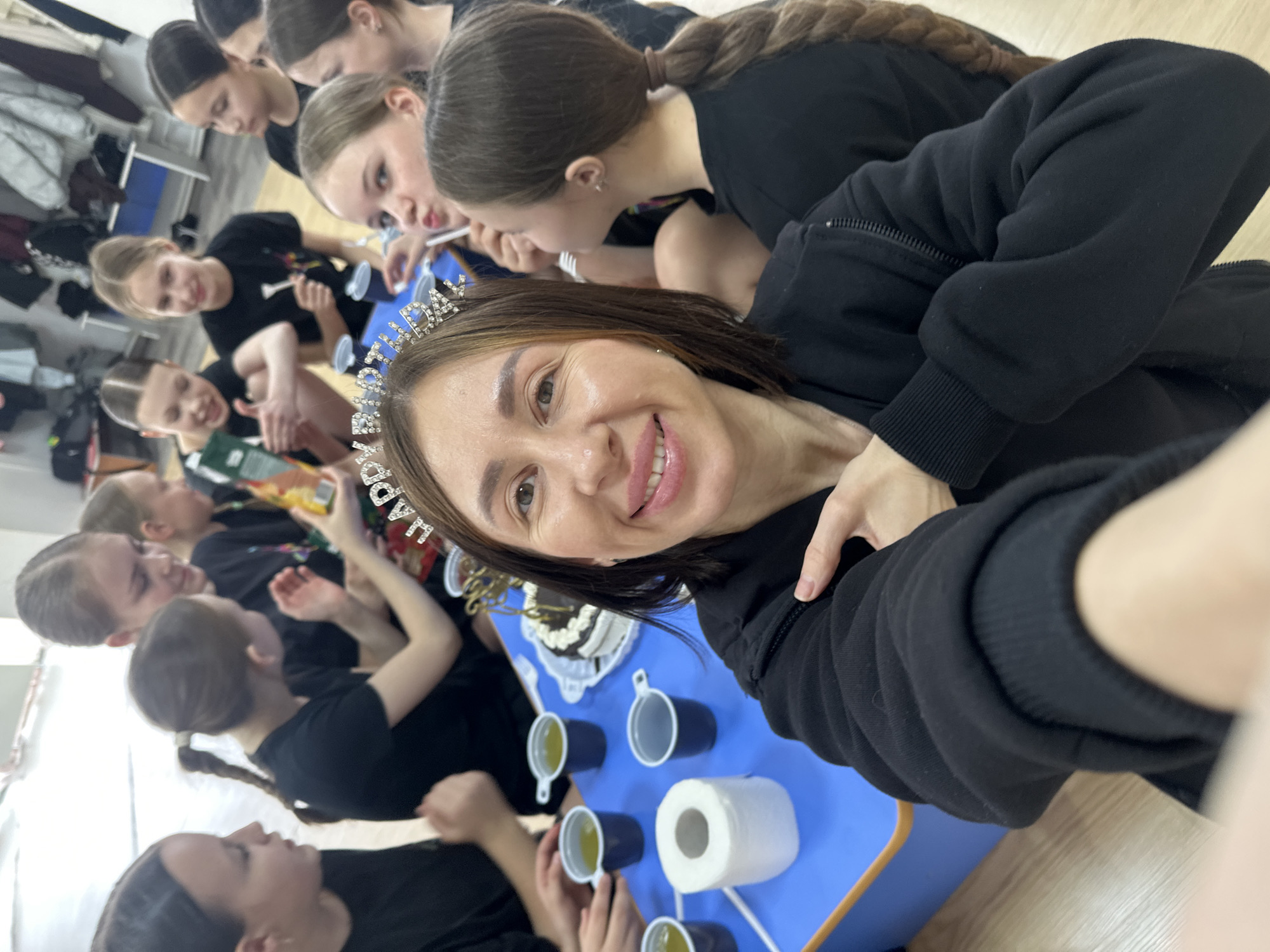 A selfie-style photograph depicts a woman with a tiara celebrating her birthday with a group of girls, likely students. The woman has shoulder-length brown hair and is wearing a black hooded jacket. She smiles directly at the camera.
Behind her, a blue table is set with cups of a light-colored liquid, a roll of toilet paper, and a cake. The girls surrounding the table have their hair pulled back, and are wearing black tops, suggesting a dance or performance context. They are engaged in the celebration, likely eating cake and interacting with each other.
The scene appears to be indoors, possibly in a dance studio or dressing room. The location might be inferred as Votkinsk, Russia, based on an external prompt. The overall ambiance suggests a birthday celebration following a performance or rehearsal.