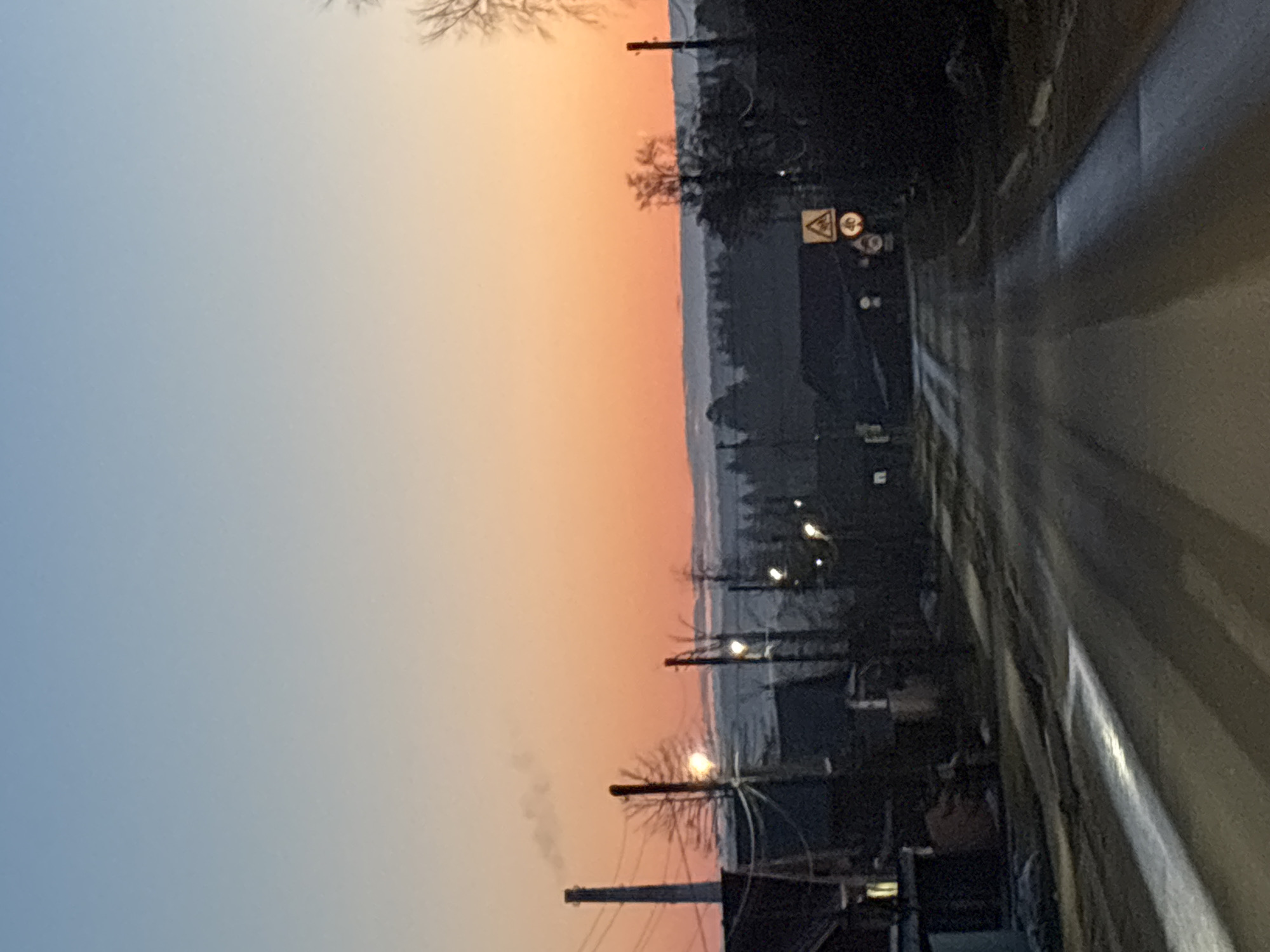 Captured at dusk in Chaikovsky, Russia, the image presents a street scene with a prominent road stretching into the distance, reflecting the fading light. The sky transitions from a soft blue to a warm orange, creating a picturesque backdrop. Along the roadside, utility poles are visible, adorned with wires and topped with lights that dot the landscape. Additionally, there are silhouettes of buildings and trees. A signpost with road warning symbols is also noticeable by the roadside.