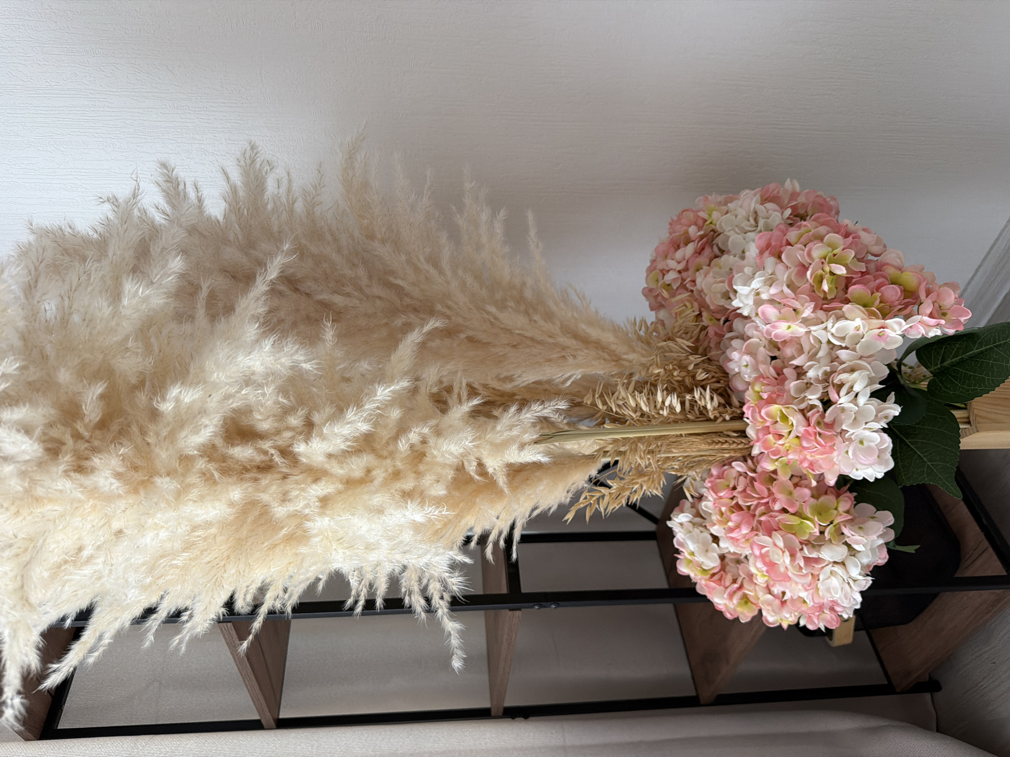 A tall, fluffy, cream-colored pampas grass arrangement dominates the center of the frame. It is set in a black pot, which is partially obscured by a cluster of pink and white hydrangea flowers at the base of the arrangement. Behind the floral arrangement, a dark metal shelving unit with three wooden shelves is visible. A white wall with a textured pattern forms the background. To the left, a sheer white curtain is partly visible. The scene appears to be an indoor setting, likely a room in a residential or commercial space. There are no people or animals present in the image. The lighting is soft and diffused, suggesting it might be daytime. The image does not contain any specific indicators that would allow for determination of its location in Moscow, Russia.