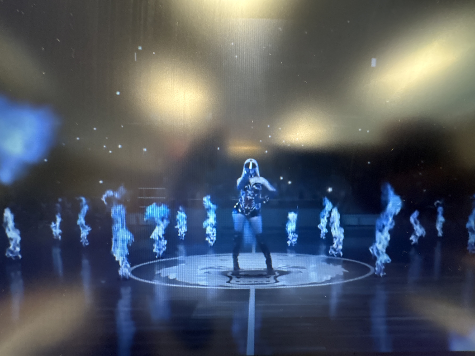 The image shows a woman in a glittering outfit and thigh-high boots standing on a circular stage. She is surrounded by ethereal blue flames. The background is dark with scattered white lights, resembling stars. The floor is reflective, showing a faint reflection of the flames and the woman. The overall scene suggests a performance or a music video. The image is a low-resolution photograph taken from a screen, possibly a television or computer monitor. There is no visible indication of the location in Sofia, Bulgaria.