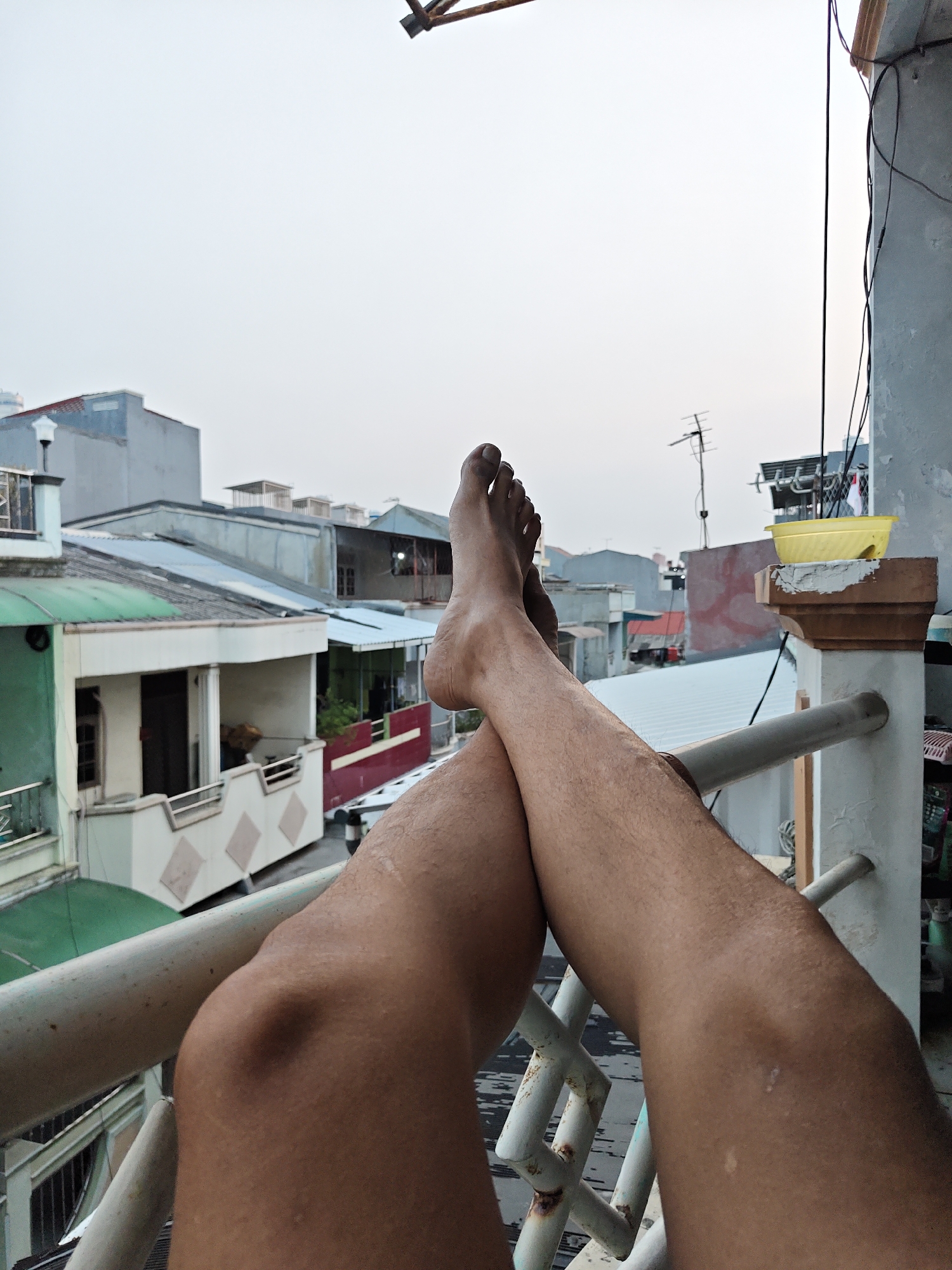 The image shows a first-person perspective of a person's legs resting on a balcony railing in North Jakarta, Indonesia. The person's feet are bare and crossed, with the toes pointing upwards. Their legs are hairy and tanned. In the background, a dense urban landscape of residential buildings with varying roof types is visible. The sky is overcast and appears to be a pale gray. The balcony railing is made of white metal with a slightly rusted appearance. The scene depicts a moment of relaxation or leisure on a balcony overlooking a city.