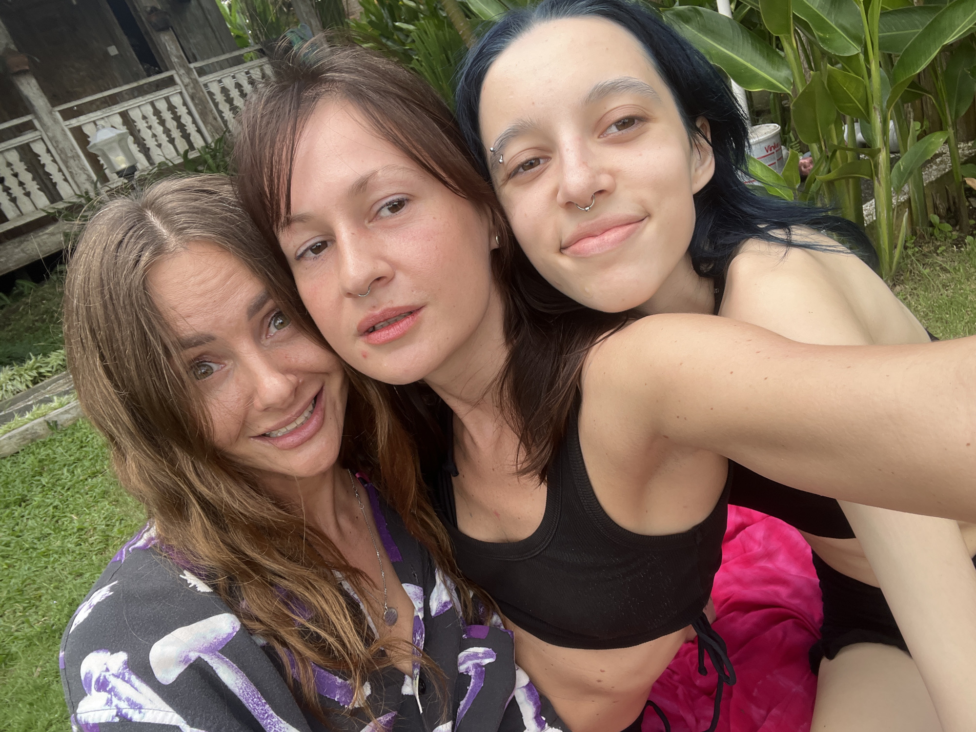 Three women are posing for a selfie outdoors. The woman on the left has long brown hair and is wearing a dark gray shirt with purple mushroom designs. The woman in the middle has medium-length brown hair and is wearing a black top. The woman on the right has dark blue hair, a piercing above her eyebrow, and a nose ring. She is wearing a black sports bra and black shorts.
Behind them is a wooden structure with decorative trim and lush green foliage, indicating they may be in a tropical location. The setting is likely within the city of Mengwi, Indonesia. A pink towel is beneath the woman on the right.