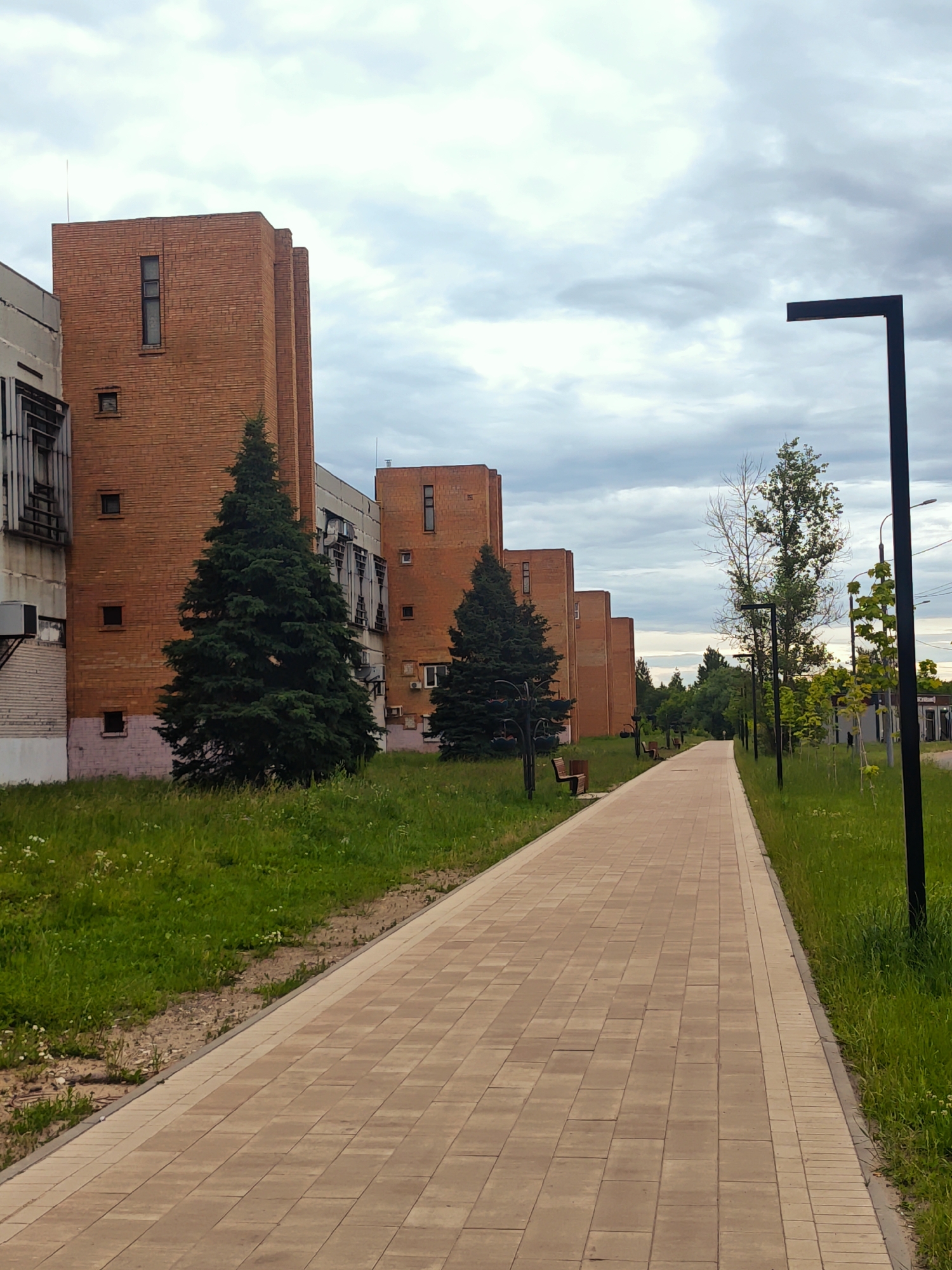 This is a vertical, eye-level, medium shot of a paved walkway lined with green grass and trees. The walkway, made of light brown rectangular pavers, stretches into the distance, curving slightly to the left. On the left side of the walkway are several brick buildings, appearing to be apartments or offices, with evenly spaced windows. Evergreen trees stand in front of the buildings, casting shadows on the grass.
On the right side of the walkway, there is more grass and several trees, including a bare-branched deciduous tree and a smaller tree with vibrant green leaves. A modern, black lamppost stands tall on the far right edge of the image, its arm extending horizontally over the walkway. Further down the walkway, another similar lamppost is visible, and beyond that, more trees and some low-lying structures suggesting a park or residential area.
The sky is overcast with soft, gray clouds, indicating a cloudy day, likely in the late afternoon or early evening, given the diffused light. The overall mood is peaceful and serene. There are no people visible in the image, but the presence of benches along the walkway suggests it's a place for recreation and relaxation. The architecture of the buildings, particularly the brickwork and window design, provides some clue about the location.