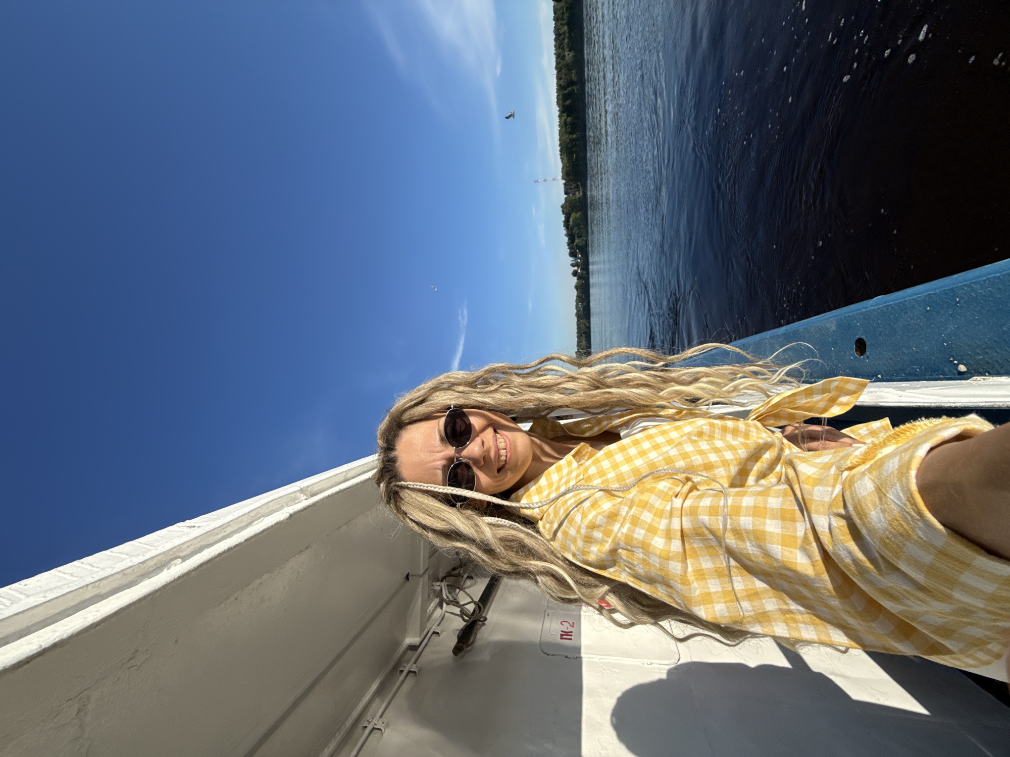 A woman with long blonde dreadlocks and dark sunglasses smiles directly at the camera, taking a selfie on a boat in the city of Dumino, Russia. She wears a yellow and white checkered button-down shirt, tied at the waist, over a white top. Her left side is against a white boat wall, which displays a red "П-2" marking and casts sharp shadows. To her right, a blue railing borders a wide expanse of dark blue water with ripples and foam. The water extends to a distant, tree-lined shore, above which a tall, slender lattice communication tower is visible. The sky above is clear blue with a few wispy clouds and two birds in flight, one appearing to be a seagull on the right side of the frame.