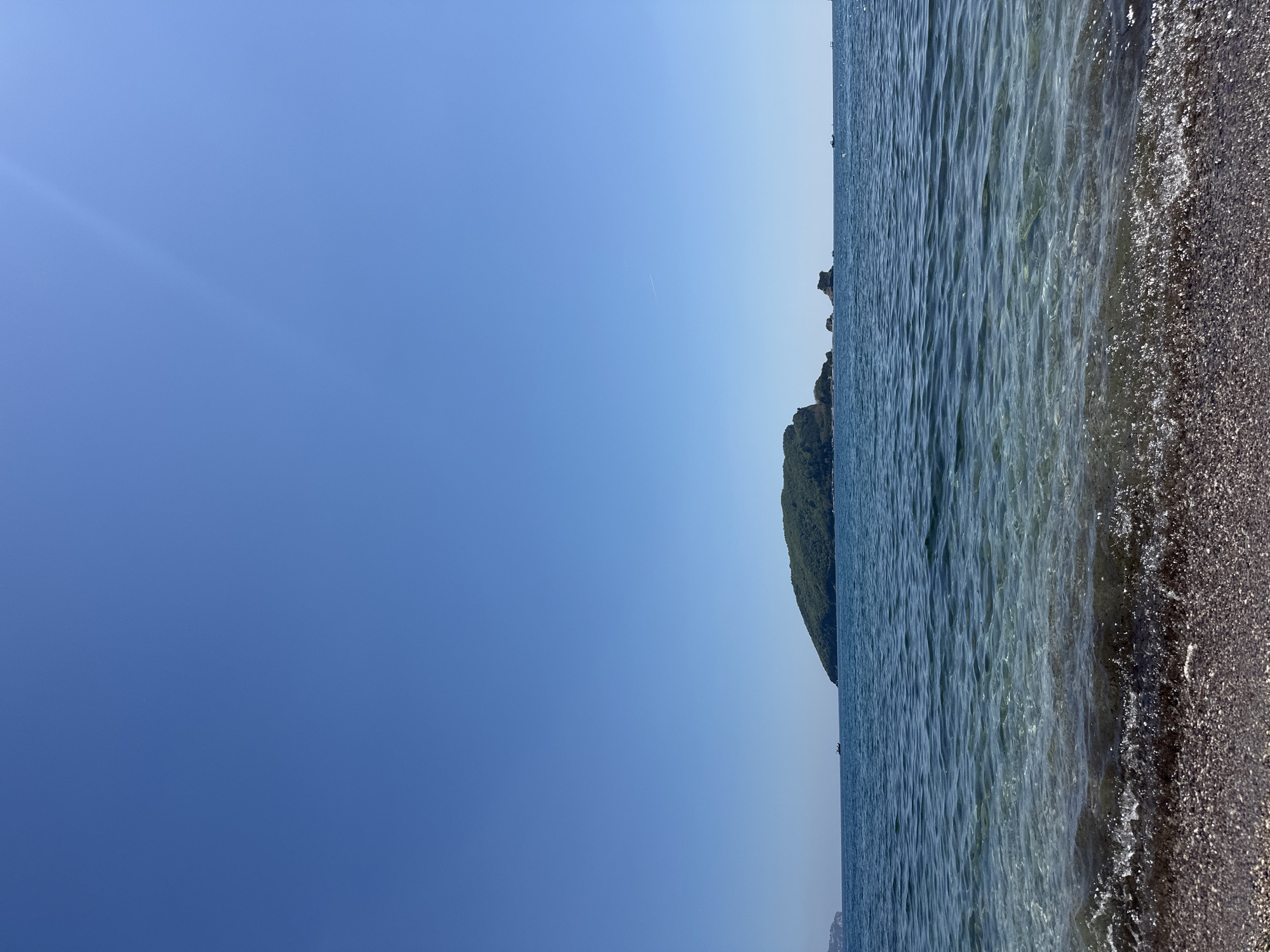 The image depicts a coastal scene, likely in Budva, Montenegro. The sky is clear and blue, with a color gradient from a deeper hue in the lower left to a lighter shade towards the right.
The right side of the image features the sea and a portion of the shoreline. The water appears calm with small waves gently lapping against the dark, pebbly beach. In the distance, a rocky, green-covered landmass can be seen.
