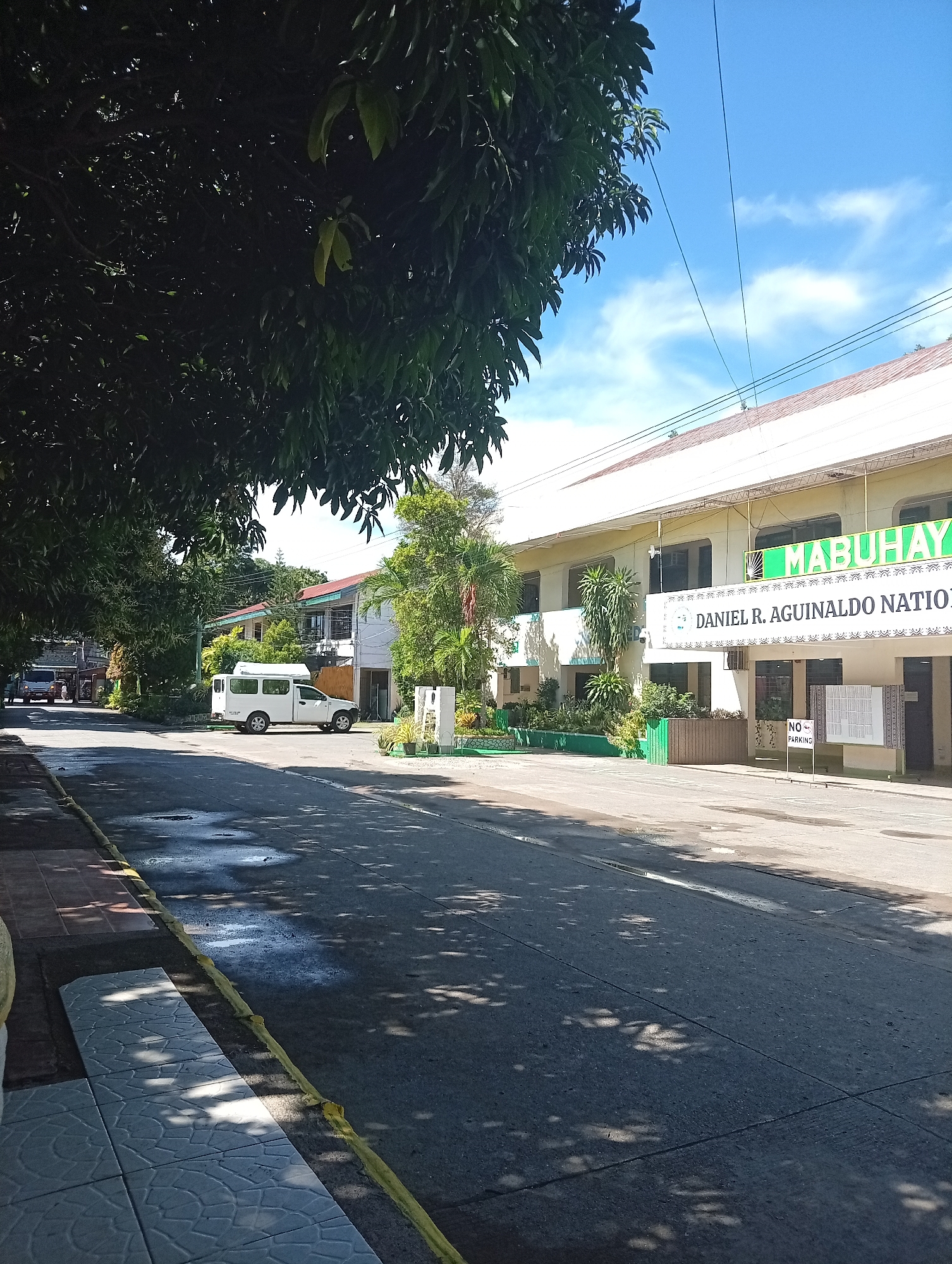 The image captures a street scene in front of Daniel R. Aguinaldo National High School in Davao City, Philippines. The foreground features a paved road with some wet patches reflecting the sky and shadows from trees. To the left, a tiled sidewalk is visible with a yellow curb along the road. In the middle ground, a white van is parked, and the school building is prominent on the right. The building has a white facade with a light-colored roof and a large banner displaying "MABUHAY" in green letters, followed by "DANIEL R. AGUINALDO NATION". A "NO PARKING" sign is placed on a stand near the entrance. Lush greenery, including trees and shrubs, surrounds the school. The sky is blue with scattered clouds. The overall scene suggests a typical day at a school in a tropical climate.