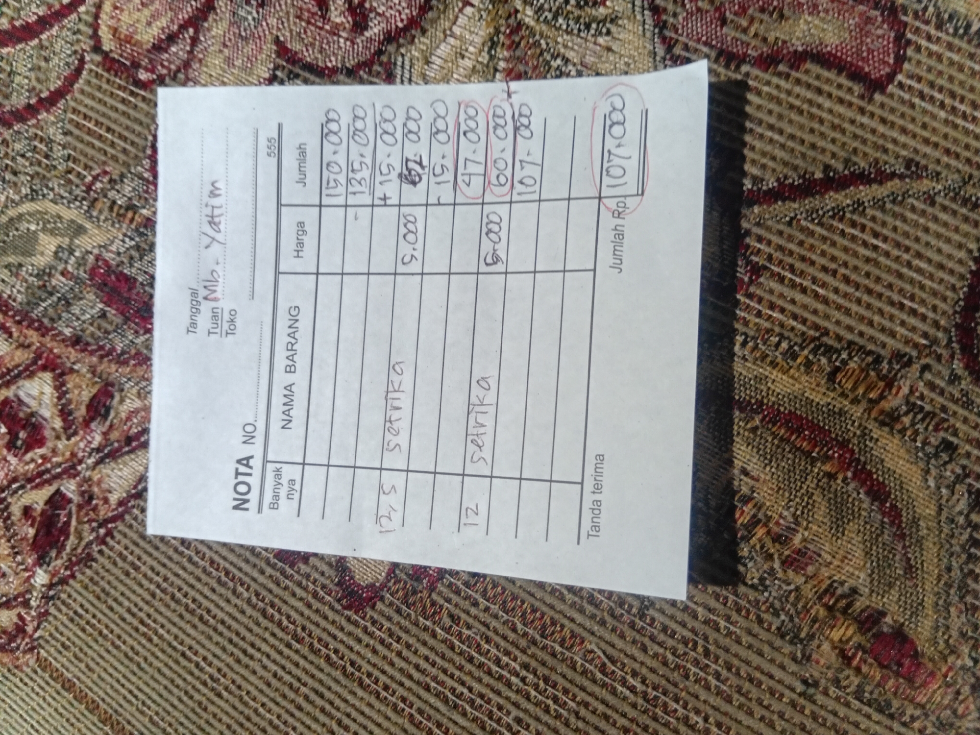 The image shows a close-up, top-down view of a handwritten receipt on a patterned fabric surface. The receipt is for "NOTA NO." and contains columns for "Banyak nya" (quantity), "NAMA BARANG" (item name), "Harga" (price), and "Jumlah" (total).
The items listed are "setrika" (iron) with quantities and prices. There are calculations written on the receipt, including subtractions and additions, leading to a final "Jumlah Rp. 107.000".
Above the item list, there is information regarding the date and customer, with "Tuan Mb. Yatim" and "Toko" written. The word "Tanda terima" (receipt) is at the bottom. The fabric surface appears to be a blanket or tablecloth with a woven, geometric pattern in shades of brown, beige, and maroon. The lighting suggests an indoor setting. There is no information to indicate the location in the city of Plemahan, Indonesia.