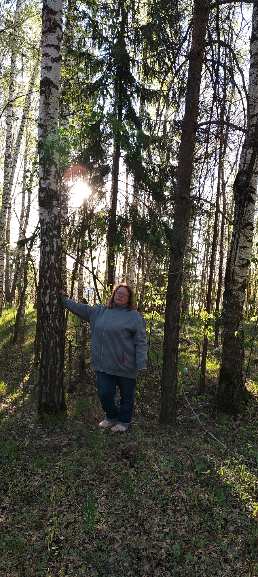 The photo was taken outdoors in a wooded area near Zipunovo, Russia. A middle-aged woman with reddish-brown hair stands in the foreground, wearing a gray zippered jacket and blue jeans. She is leaning against a birch tree on the left side of the frame. The background consists of a forest with various trees, including birch and evergreens. Sunlight filters through the trees. The ground is covered in leaves and some green vegetation.