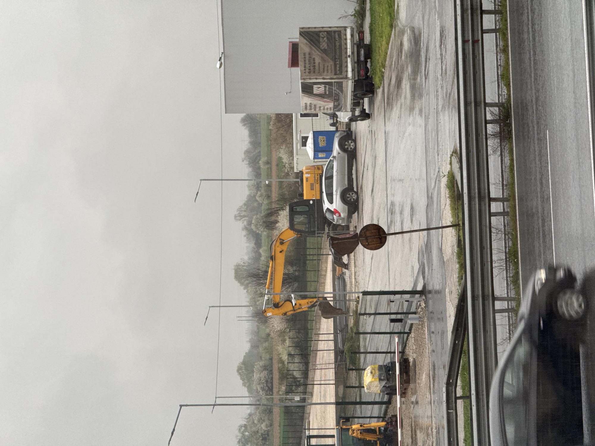 The image shows a roadside scene in Unknown, Bulgaria, on a cloudy, rainy day. An excavator is visible behind a fence, along with various parked vehicles and construction equipment. A road runs alongside the scene, with a car in motion and a metal safety barrier. Overhead lighting is present, suggesting an industrial or commercial area.