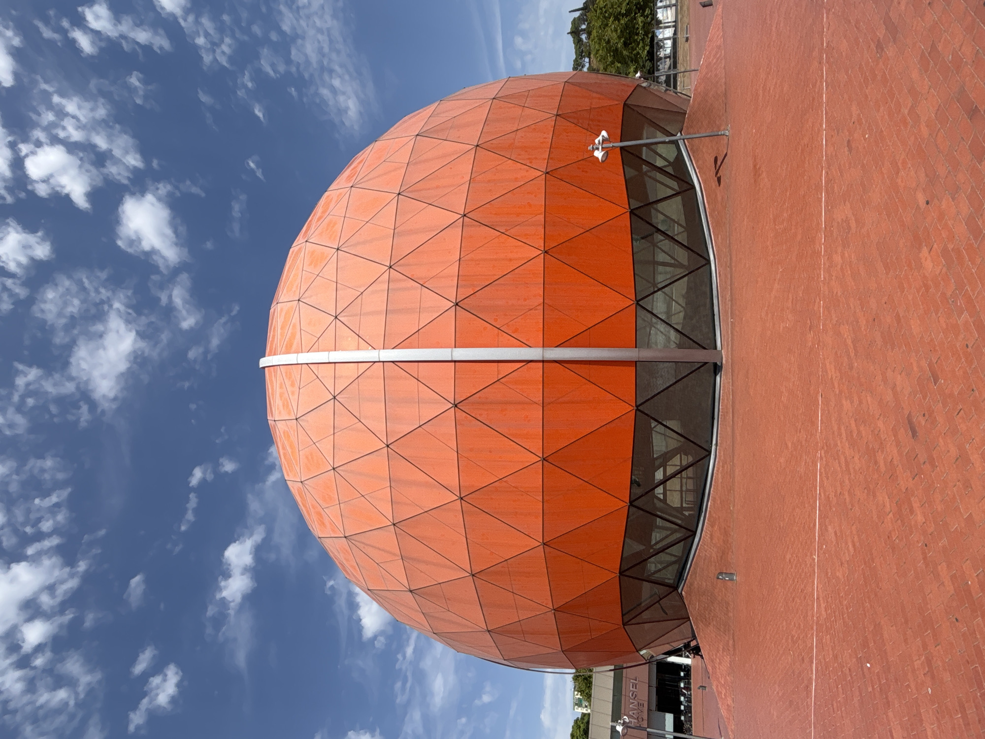 This is a full-frame, eye-level, vertical, outdoor, daytime, low-angle shot of the iconic Orange Sphere (La Badalona Sphere), a large geodesic dome structure located in Badalona, Spain. The sphere is predominantly bright orange and is covered in a pattern of triangular panels. A prominent vertical silver pole runs down the center of the sphere. The lower section of the sphere features a band of reflective glass windows with a geometric metal framework visible behind them.
The sphere is situated on a wide, flat plaza paved with red brick. The plaza is clean and empty except for a few small bollards and two modern-looking streetlights. In the background, to the left of the sphere, is a building with signage that reads "HANSEL HOME". Further in the distance, trees and buildings are visible. The sky above is a vibrant blue, scattered with fluffy white clouds. The lighting is bright, indicating a sunny day.