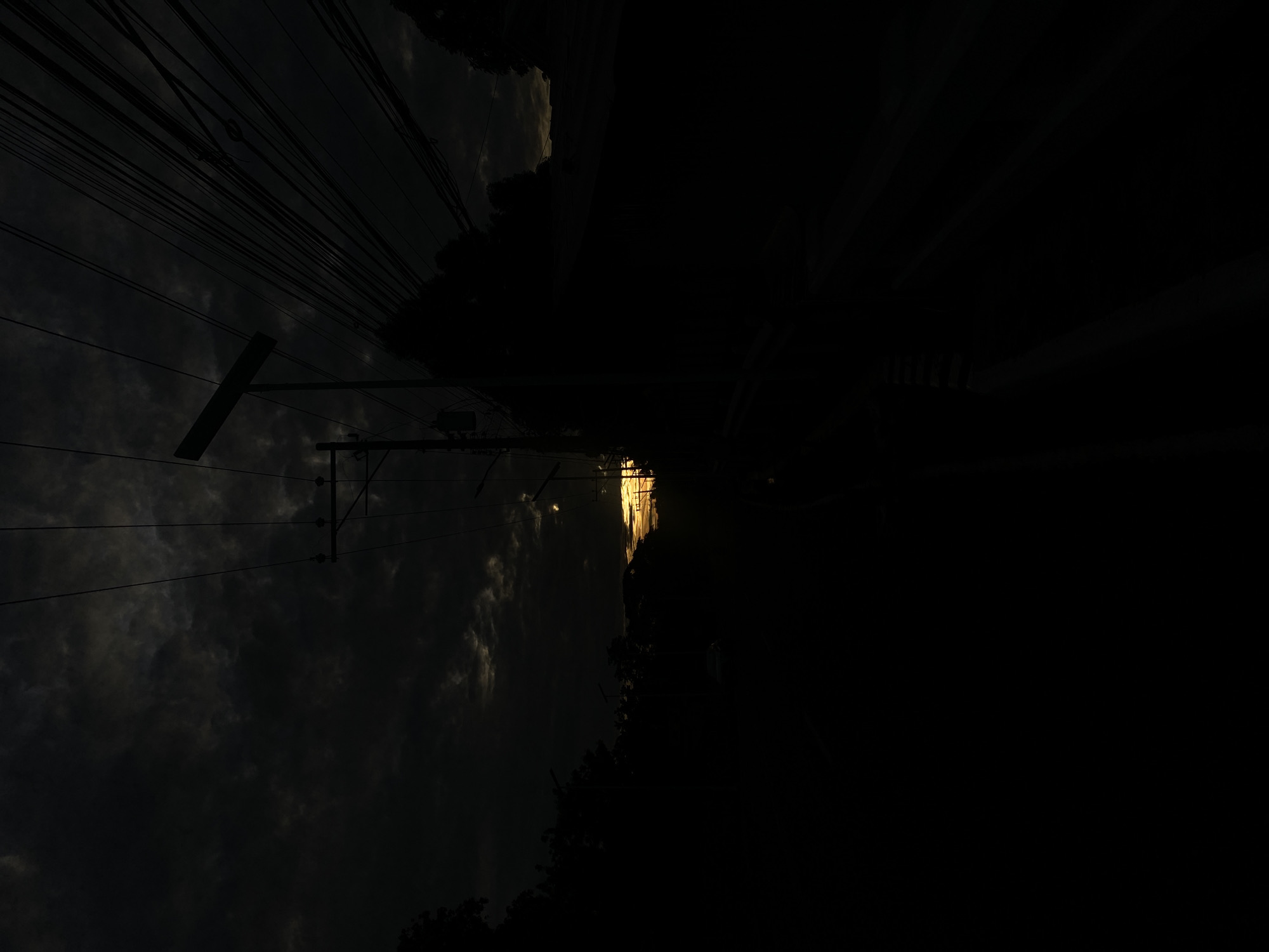 The image captures a dramatic twilight scene in Davao City, Philippines, likely near a roadside with overhead power lines. The sky is dominated by dark, ominous clouds, with a sliver of bright, orange-yellow light piercing through near the horizon, suggesting a sunset or sunrise. Silhouetted against this dramatic sky are the stark shapes of utility poles and a dense network of electrical wires. Trees and other vegetation are also visible in silhouette along the sides of what appears to be a road, obscuring much of the foreground and background details. The overall impression is one of encroaching darkness, with the natural light struggling to break through the heavy cloud cover.