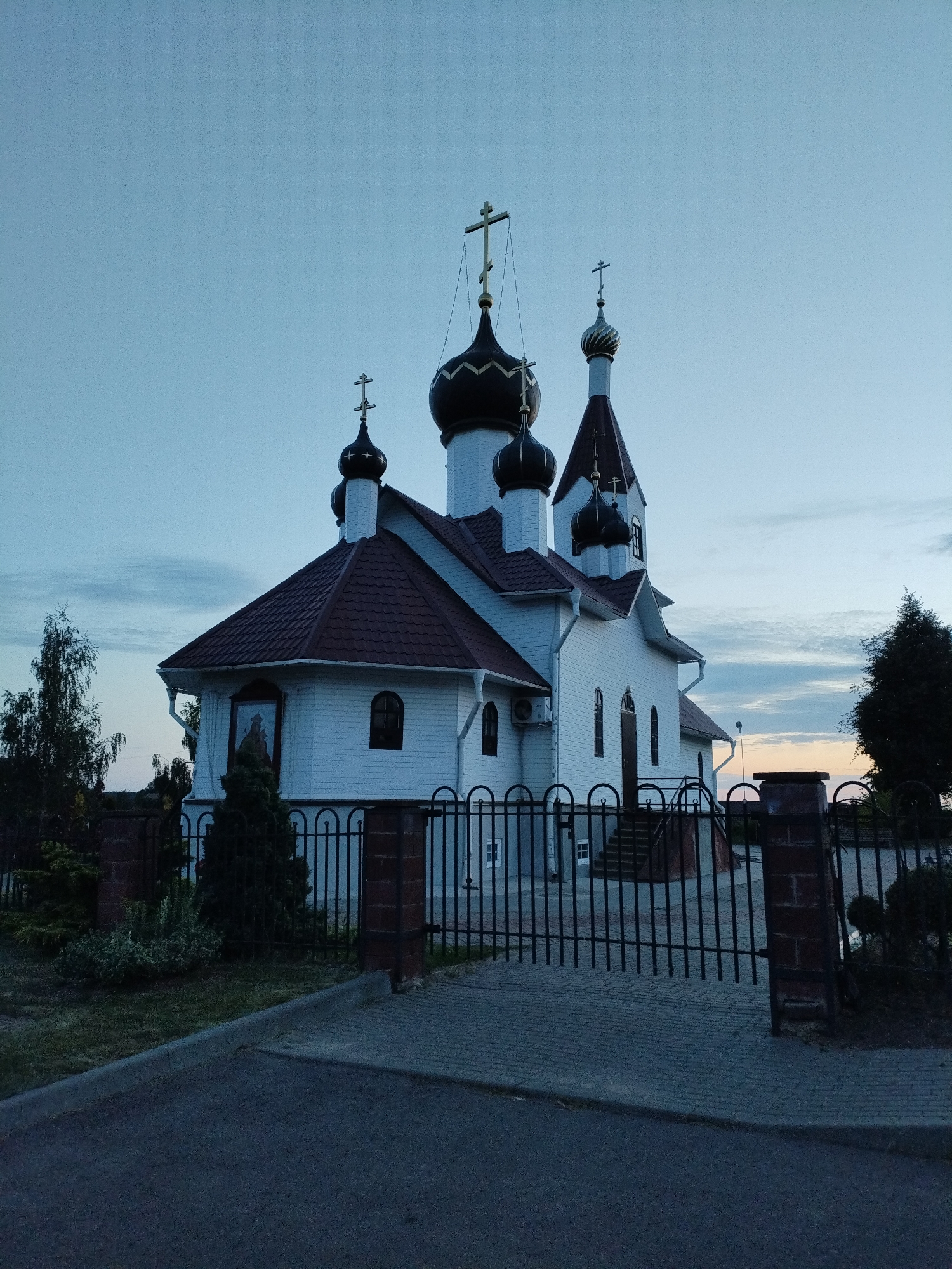 This image shows a white wooden church with a dark tiled roof and dark onion domes, each topped with a cross. The church is located in the city of Bialynichy, Belarus. The church is fenced with a black metal gate and brick pillars. The sky is a pale blue with some clouds, and it appears to be dusk or early morning. There are trees and bushes around the church. The ground in front of the church is paved with cobblestones and asphalt. There are no people visible in the image.