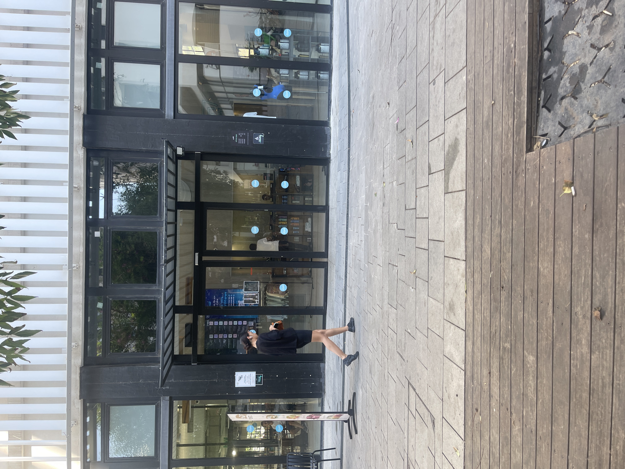 A medium shot shows a woman walking on a paved sidewalk in front of a modern building with large glass windows. The woman, wearing a black shirt and shorts, is walking from left to right, facing slightly away from the camera. She has earbuds in and is holding a brown bag in her right hand. The building features a black facade with multiple large windows. Some windows display merchandise, while others reflect the surroundings. A few people are visible inside the building through the glass. The sidewalk is made of light-colored rectangular tiles, and in the foreground, there is a wooden deck area. The overall scene appears to be in a commercial or public area of Tel Aviv, Israel.