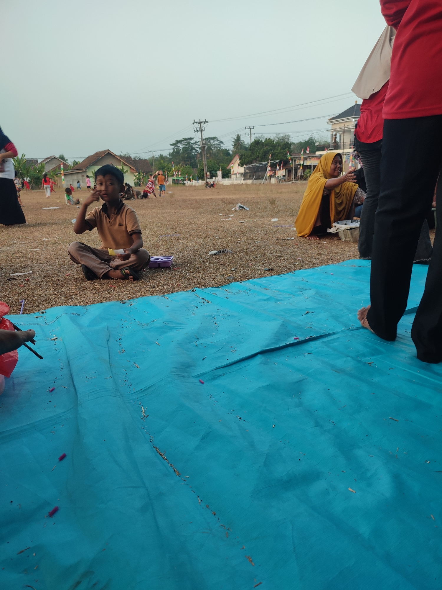 In Nusa Jaya, Indonesia, on a dry, open field with scattered dry grass and debris, a group of people are gathered. In the foreground, a young boy in a tan polo shirt and brown pants sits cross-legged on the ground, holding a small yellow object and touching his face. A large, bright blue tarp covers a significant portion of the foreground, with some small red objects and dry plant matter scattered on it. To the right of the boy, a woman wearing a yellow hijab and dark clothing is partially visible, looking to her right and reaching out. Behind them, several other individuals are present, including a person in a red shirt and black pants on the far right, with bare feet visible. In the background, there are houses with brown roofs, trees, and utility poles. The sky is overcast and pale. The overall scene suggests an outdoor gathering or event.