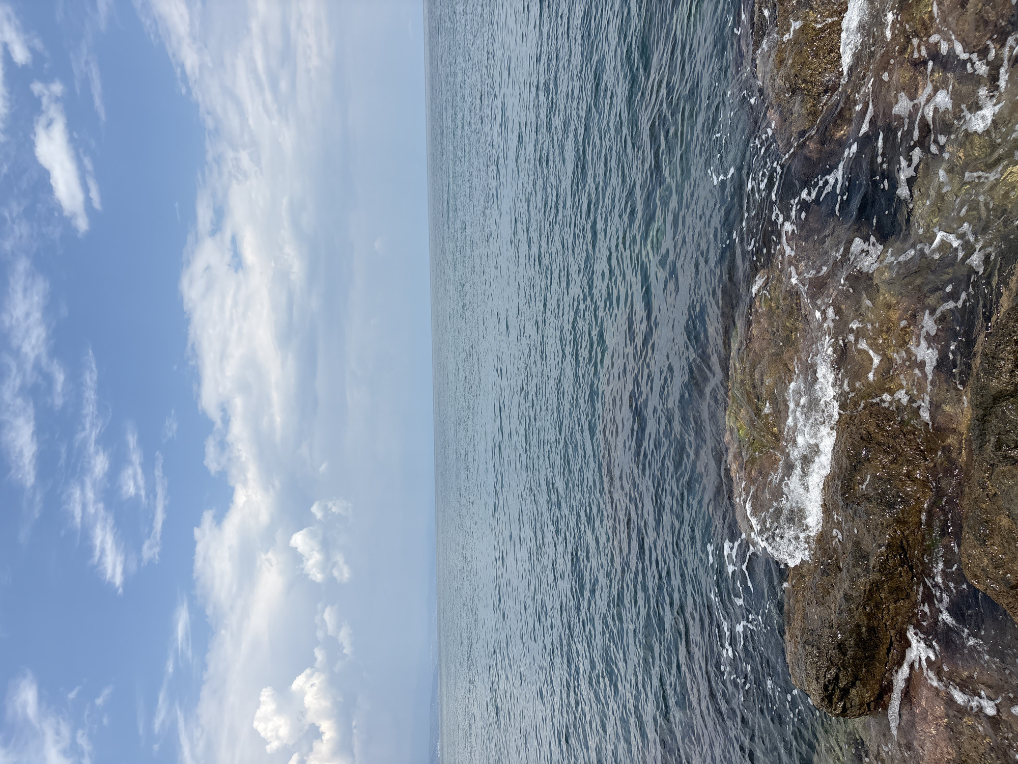 The image shows a body of water, likely the sea or a large lake, with a rocky shoreline in the foreground. The water is dark blue with ripples and some white foam from waves breaking on the rocks. The rocks are varied in color and texture, with some appearing wet and dark, while others are lighter brown. The sky above is partly cloudy, with patches of blue sky visible between white and gray clouds. In the distance, a faint outline of land or mountains can be seen on the horizon. There are no people or animals in the image. The scene is a natural landscape, likely depicting a coastal or lakeside environment.