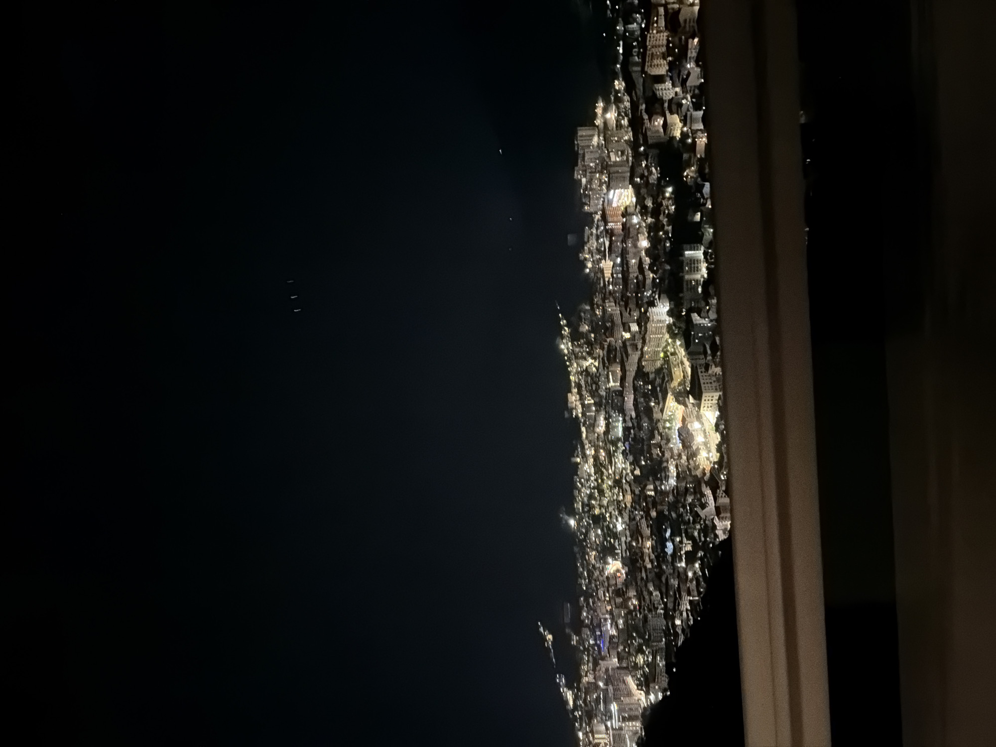 The image shows a nighttime view from a high vantage point, likely a window, overlooking the city of Budva, Montenegro. The right side of the image shows city lights stretching along the coastline. To the left is the dark expanse of the Adriatic Sea. In the far distance, three faint lights are visible on the water. The scene is viewed through a window frame, with the right portion of the frame and curtains slightly visible. The city appears dense with a mix of buildings and residential areas illuminated by streetlights, car lights, and building lights. The photograph seems to have been taken at an angle.