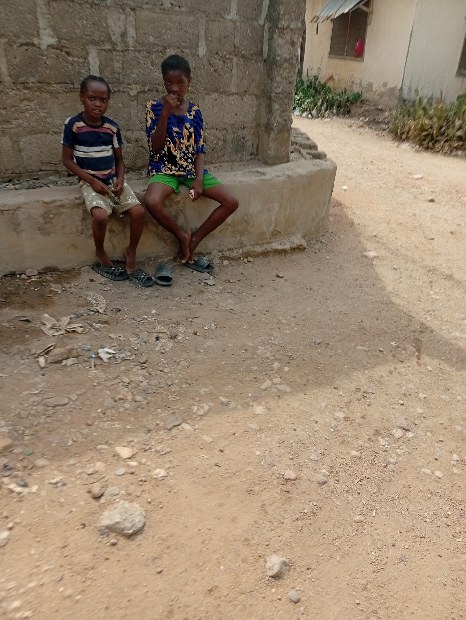 Two children are seated on a low, weathered concrete ledge in what appears to be a rural or semi-urban setting in Adankolo, Nigeria. The child on the left, wearing a striped t-shirt and light-colored shorts, is looking directly at the camera, holding a small object in their hands. The child on the right, dressed in a blue patterned long-sleeve top and green shorts, also looks towards the viewer, with one hand raised near their mouth.
Behind the children is a rough concrete block wall. The ground in front and to the right of the ledge is dry, light brown dirt, strewn with small rocks and some discarded items like plastic bags and paper. Several pairs of dark slippers or sandals are placed on the dirt beneath where the children are sitting.
In the background to the right, a dirt path or road leads past simple, light-colored buildings with basic windows and corrugated metal roofs. Patches of green vegetation are visible around the base of these structures. The scene is brightly lit by natural daylight, indicating clear weather. No visible text is present in the image.