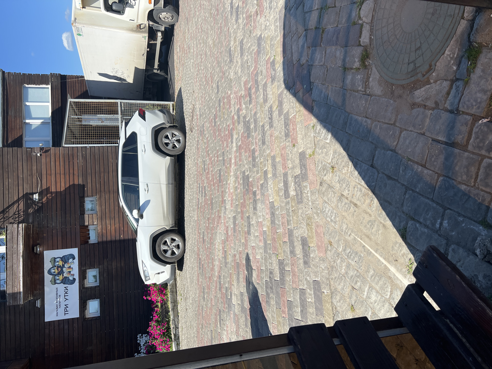In Gavrilkovo, Russia, a white SUV is parked on a cobblestone street in front of a wooden building. The building has a sign that reads "МАГАЗИН РАЗЛИВНЫХ НАПИТКОВ" and "ТРИ УТКИ," indicating a store selling draft beverages with the name "Three Ducks." To the right of the SUV, a white truck is partially visible. The street is paved with colorful cobblestones, and a shadow of the SUV falls across them. Pink and white flowers are planted in a bed near the building. A dark wooden bench is positioned in the foreground, partially obscuring the view. The sky is blue with some scattered clouds.