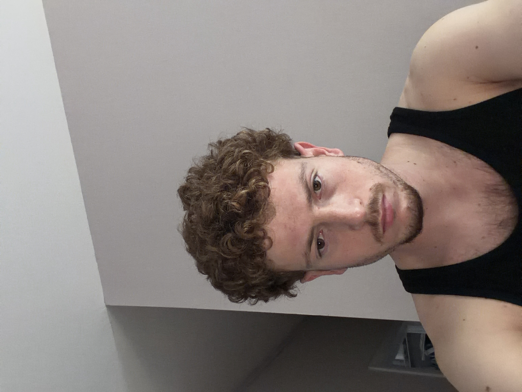 A head-and-shoulders shot of a Caucasian male with curly brown hair, brown eyes, and light skin, wearing a black tank top. The background is a plain white or light gray wall and ceiling. The location cannot be determined from the information given.
