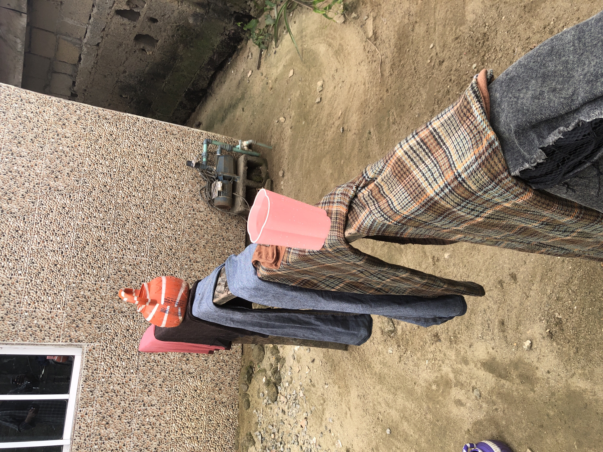 In Ado, Nigeria, an outdoor scene unfolds with laundry drying on a wooden railing. The railing, supported by posts, is laden with various items of clothing. To the left, a pink garment is draped, followed by a dark brown item, then a pair of blue jeans, and finally a plaid fabric with orange and brown hues. A pink plastic cup, partially filled with a liquid, rests on the railing, juxtaposed against the plaid clothing. In the background, a textured stone wall of a building creates a rustic backdrop. To the right of the wall, a water pump sits on concrete blocks, connected to pipes. The ground is a mix of dirt and small stones, indicative of an unpaved area. A partially visible purple object lies on the ground to the far left. The overall impression is one of everyday life in an outdoor setting.