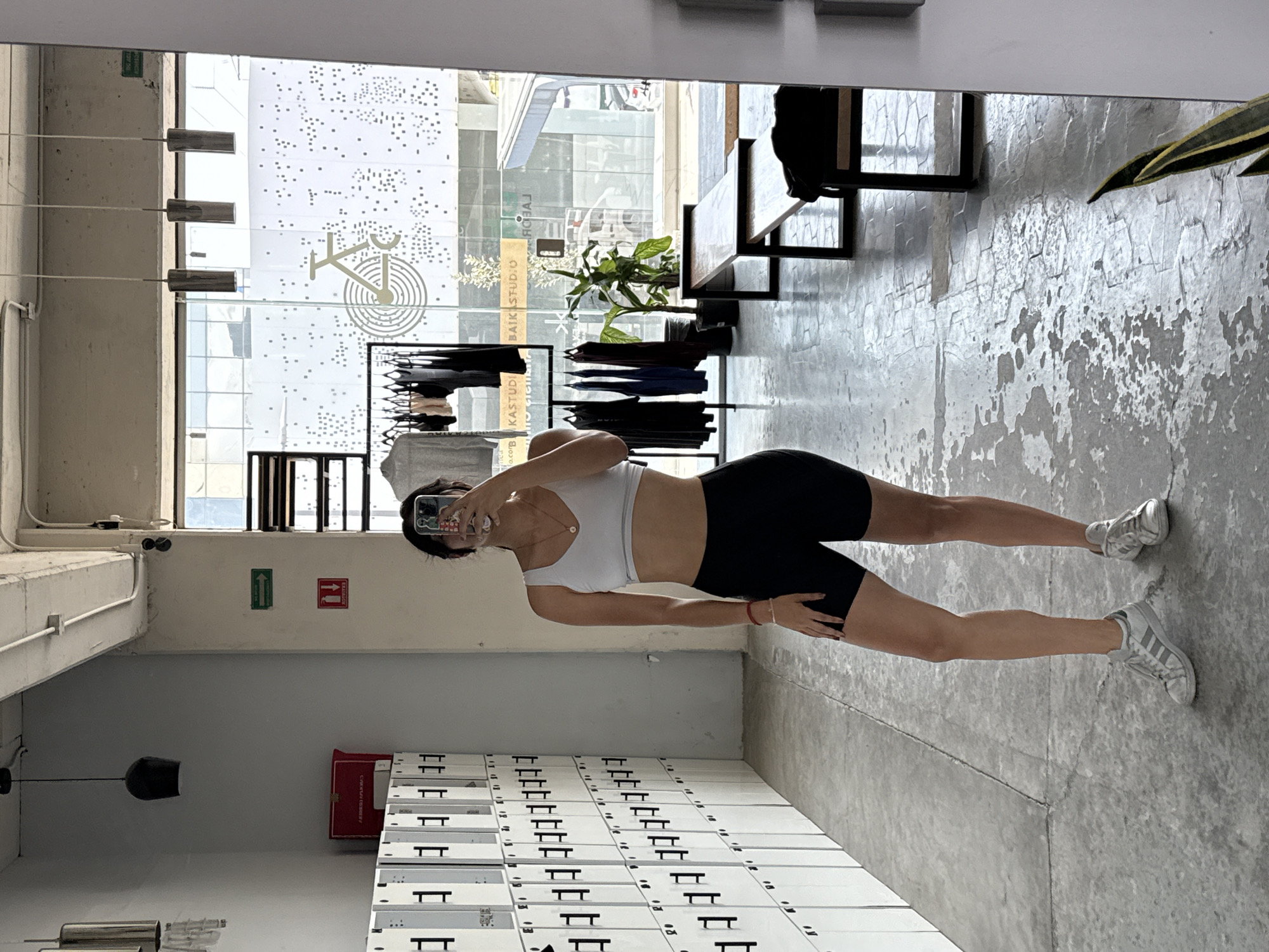 A woman stands in a mirrored gym space, holding up her phone to take a picture of herself. She's wearing a white sports bra and black shorts. The gym features a row of white lockers against one wall. Behind her, through a large window, is a street scene with the words "BAIKSTUDIO" visible on a sign, indicating a location in Puebla, Mexico. The gym itself contains exercise equipment and furniture.