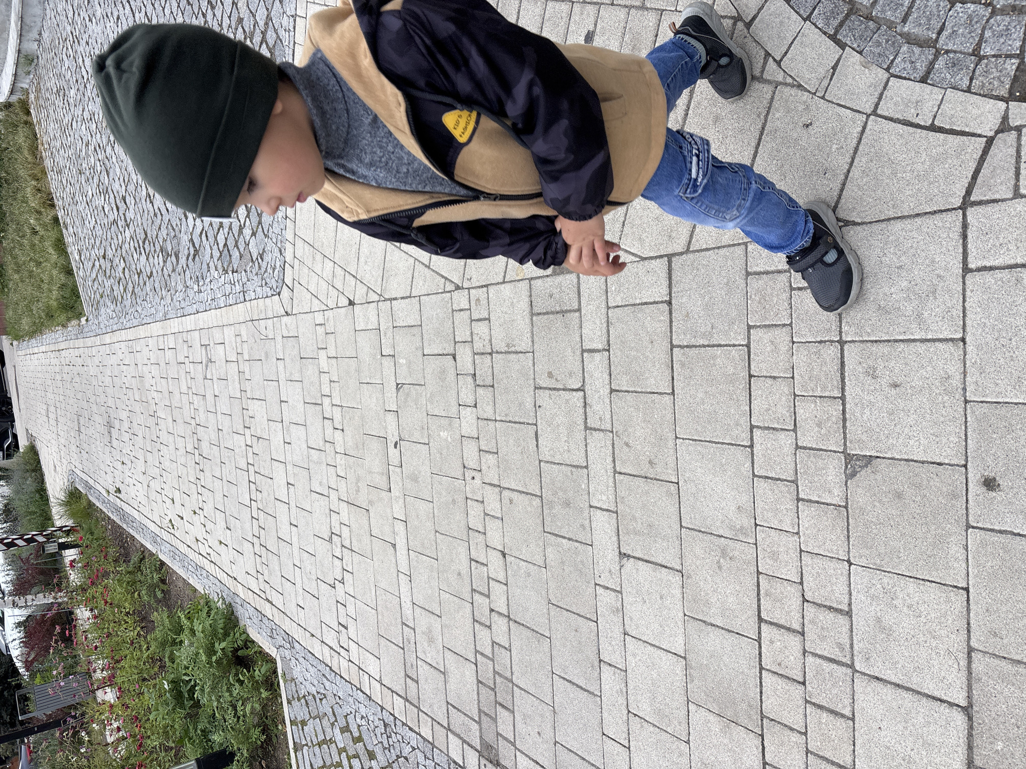 A young child, wearing a dark green beanie, a grey turtleneck, a tan and black jacket with camouflage sleeves, and blue jeans, walks on a paved walkway. The child is viewed from the chest down, in profile, looking to the right of the frame. The walkway is constructed of light grey rectangular pavers arranged in a pattern, with a border of smaller grey pavers. To the left of the walkway, there is a garden bed with green foliage and red flowers, and a metal trash receptacle. In the background, a portion of a white car is visible, as well as trees and buildings. The scene appears to be in a public outdoor space, possibly a park or a street in Zvenigorod, Russia, given the context provided. The lighting suggests it is daytime.