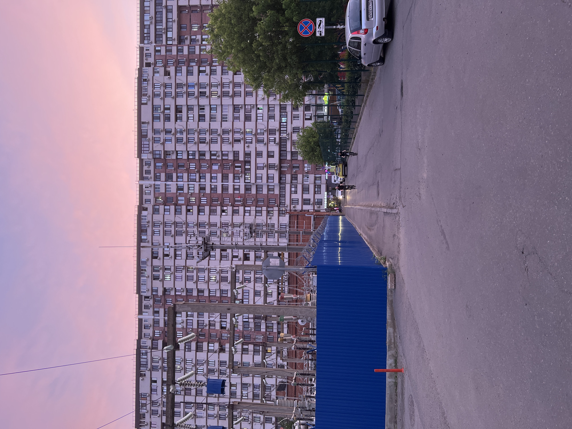 A street in Krasnodar, Russia features a high-rise apartment building with numerous windows and air conditioning units jutting out. In the foreground, an electrical substation is visible, shielded by a blue corrugated fence. A grey car is parked along the roadside, next to a "No Parking" sign. Two people on scooters are seen in the distance. The sky overhead is a gradient of colors, transitioning from pink to purple, indicating either sunrise or sunset. The overall scene is an urban setting with a mix of residential and industrial elements.