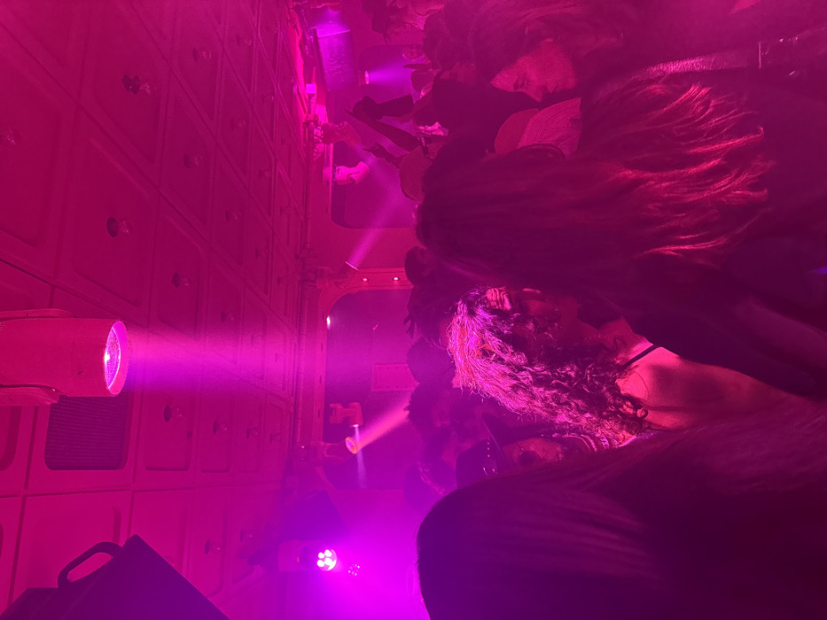 A crowd of people are in a room illuminated by pink light. A wall with cubbyholes is on the left with a light fixture projecting a pink beam. The scene is blurry and indistinct, possibly due to the low light and movement of the people. Many faces are obscured and only the outlines of bodies are visible.