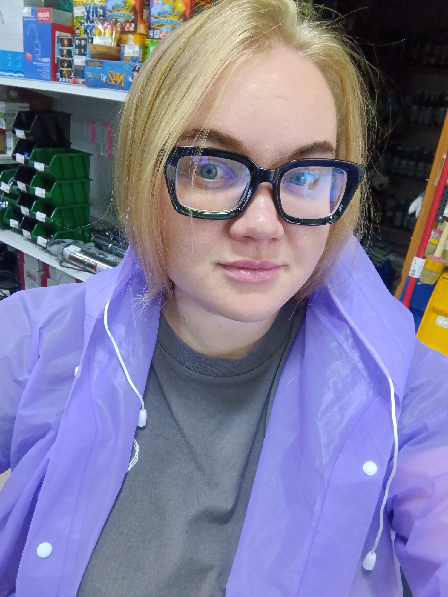 A woman with blonde hair and glasses is taking a selfie indoors. She is wearing a gray t-shirt and a light purple raincoat. The background shows shelves stocked with various items, possibly in a store or warehouse. The image was taken in the city of Foky, Russia.