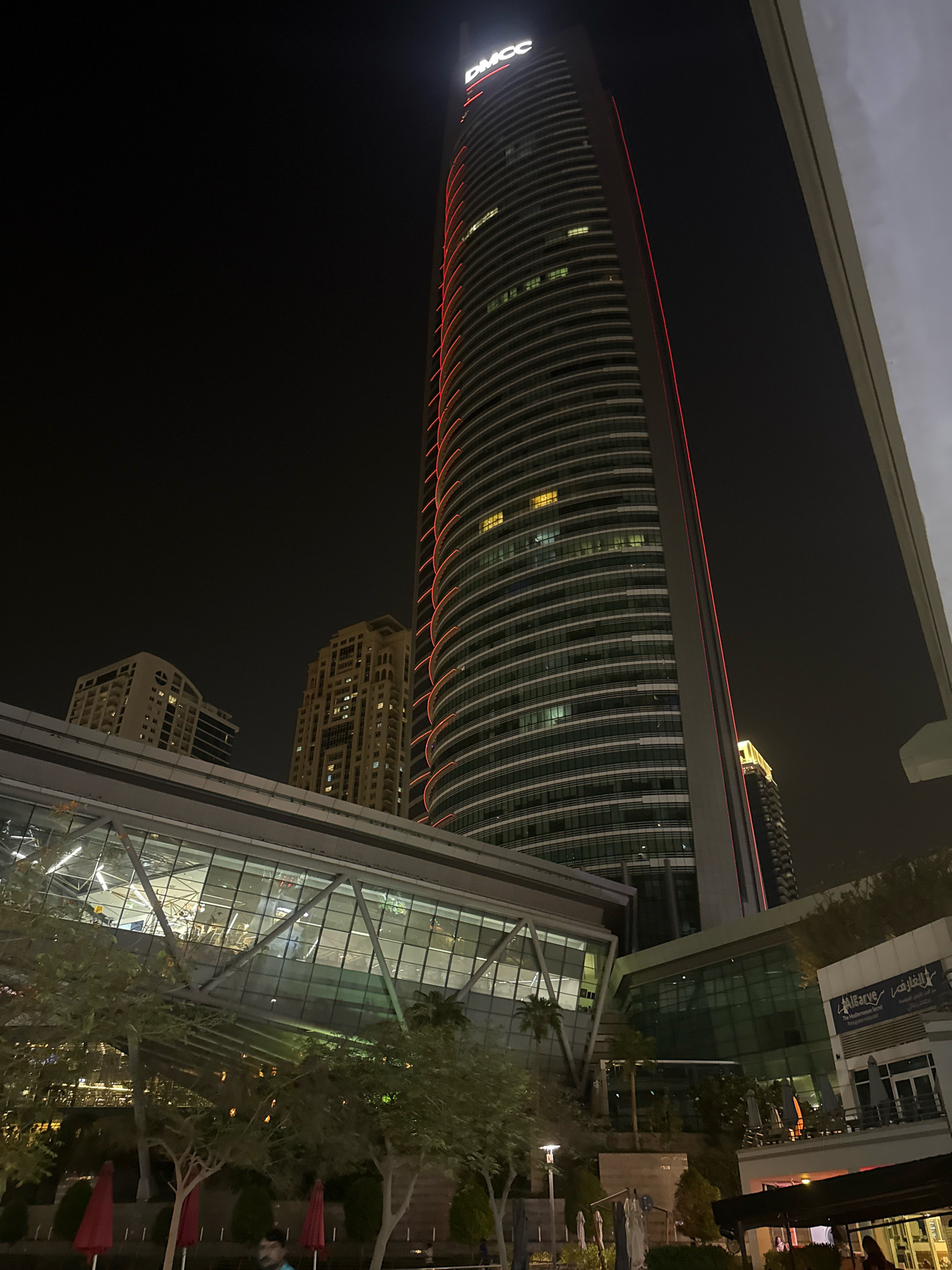 The image captures a night scene in Dubai, United Arab Emirates, likely at the Jumeirah Lakes Towers (JLT) area, given the presence of the DMCC skyscraper. The DMCC building, a prominent skyscraper, is illuminated with red vertical accents along its facade, highlighting its modern architecture.
In the foreground, a contemporary architectural structure with a glass-enclosed walkway is visible, extending across the scene. Trees with sparse foliage and some red umbrellas are scattered around the lower level. The walkway bridges over an area that appears to be an outdoor dining or seating space, with tables and chairs under umbrellas.
Behind the DMCC building, other skyscrapers of varying heights and architectural styles are visible in the background, also showing illuminated windows. One building to the left of DMCC is lighter in color and has a more geometric design. Another, to the right and further back, is darker and also appears to be a skyscraper. The sky is dark, indicating nighttime.
Several people are present in the lower part of the image, some appearing to be walking or standing in the outdoor area. One person is partially visible in the lower left corner. The lighting is a mix of artificial illumination from the buildings, the walkway, and streetlights, creating a vibrant urban atmosphere. The overall impression is one of a modern, bustling commercial and residential district at night.