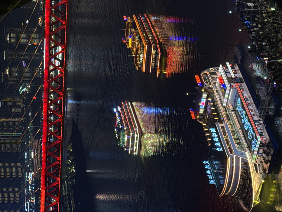 This night-time image depicts a bustling river scene within the Yuzhong District of Chongqing, China. The primary subjects are three multi-deck river cruise ships illuminated with a dense array of colorful LED lights.
In the lower foreground, a large, modern cruise ship with a white hull and multiple decks is docked at a pier on the river's edge. Its exterior is outlined with yellow-white lights, and blue Chinese characters are prominently displayed on its upper deck structure. A crowd of people is visible on the pier, some appearing to embark or disembark the vessel, while others gather along the waterfront.
In the midground, two additional illuminated cruise ships navigate the dark river. The vessel closer to the viewer features traditional architectural elements on its upper decks, including ornate roofs, and is adorned with red, yellow, and white lights. The other vessel, positioned further upriver, has a more contemporary design with green and white lights outlining its structure. Both moving ships create distinct reflections of their colorful lights on the water's surface.
Spanning the upper portion of the frame is a large, red steel truss bridge, identified as the Dongshuimen Bridge. The bridge structure itself is brightly illuminated in red, with additional smaller lights visible along its framework. Numerous pedestrians are visible walking on the bridge's upper level.
In the distant background, beyond the bridge, the dense urban landscape of Yuzhong District rises, characterized by numerous high-rise buildings displaying arrays of white and yellow lights, indicative of an active city at night. The river's surface is dark, broken only by the shimmering reflections of the boats and distant city lights.