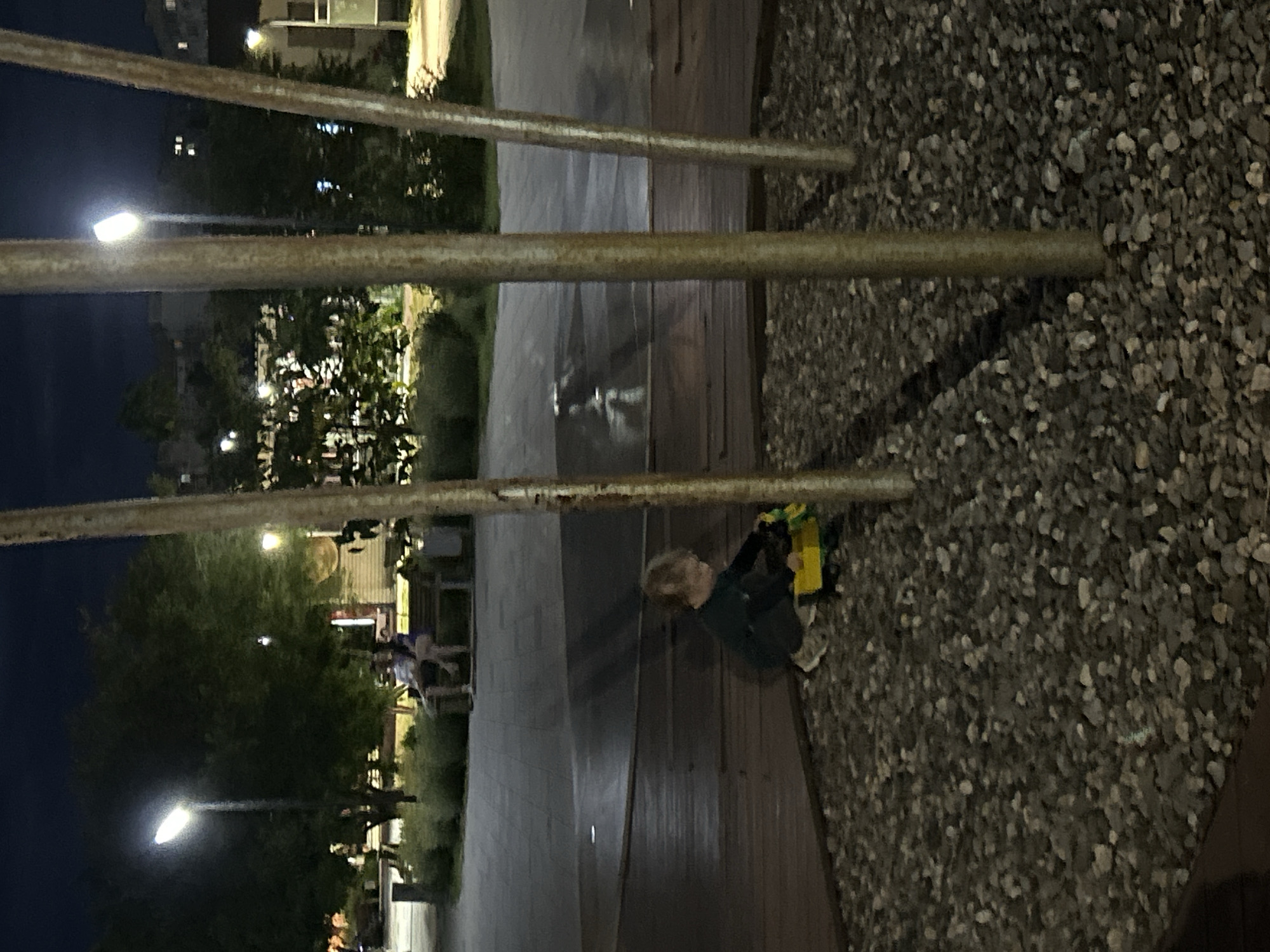 The image captures an outdoor scene at dusk. In the foreground, a young child is sitting on a wooden platform and playing with a yellow toy truck. The child is wearing a dark green jacket and grey sneakers. The platform is bordered by a gravel area on the right. In the background, there are trees, streetlights, and buildings, suggesting an urban or suburban setting. The sky is dark blue, indicating it is either early evening or late night. The overall mood is quiet and peaceful, with the child engrossed in play. There is no visible text in the image. The image appears to be taken in a public space, possibly a park or a residential area, given the presence of streetlights and buildings. The weather appears to be clear.