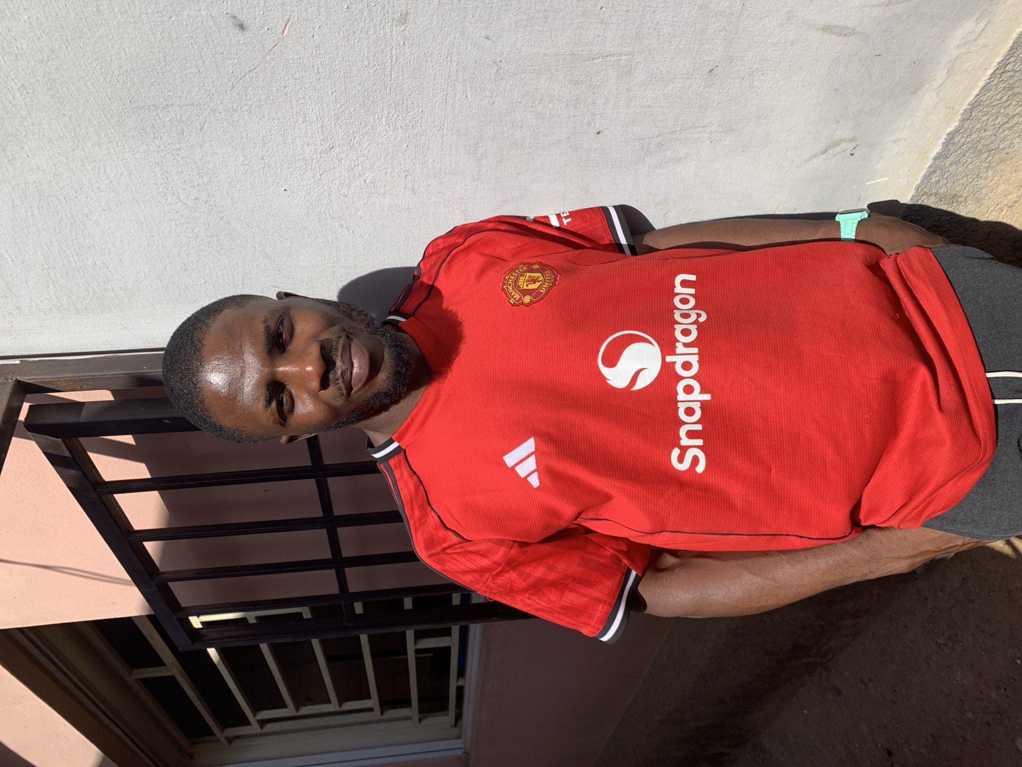 A man is wearing a red Manchester United soccer jersey with "Snapdragon" printed on the front. The jersey has the Adidas logo on one shoulder. He is also wearing a light green watch on his wrist. He is standing outside in a location near New Stadium, Nigeria, next to a wall and a metal railing. The lighting suggests it is daytime.