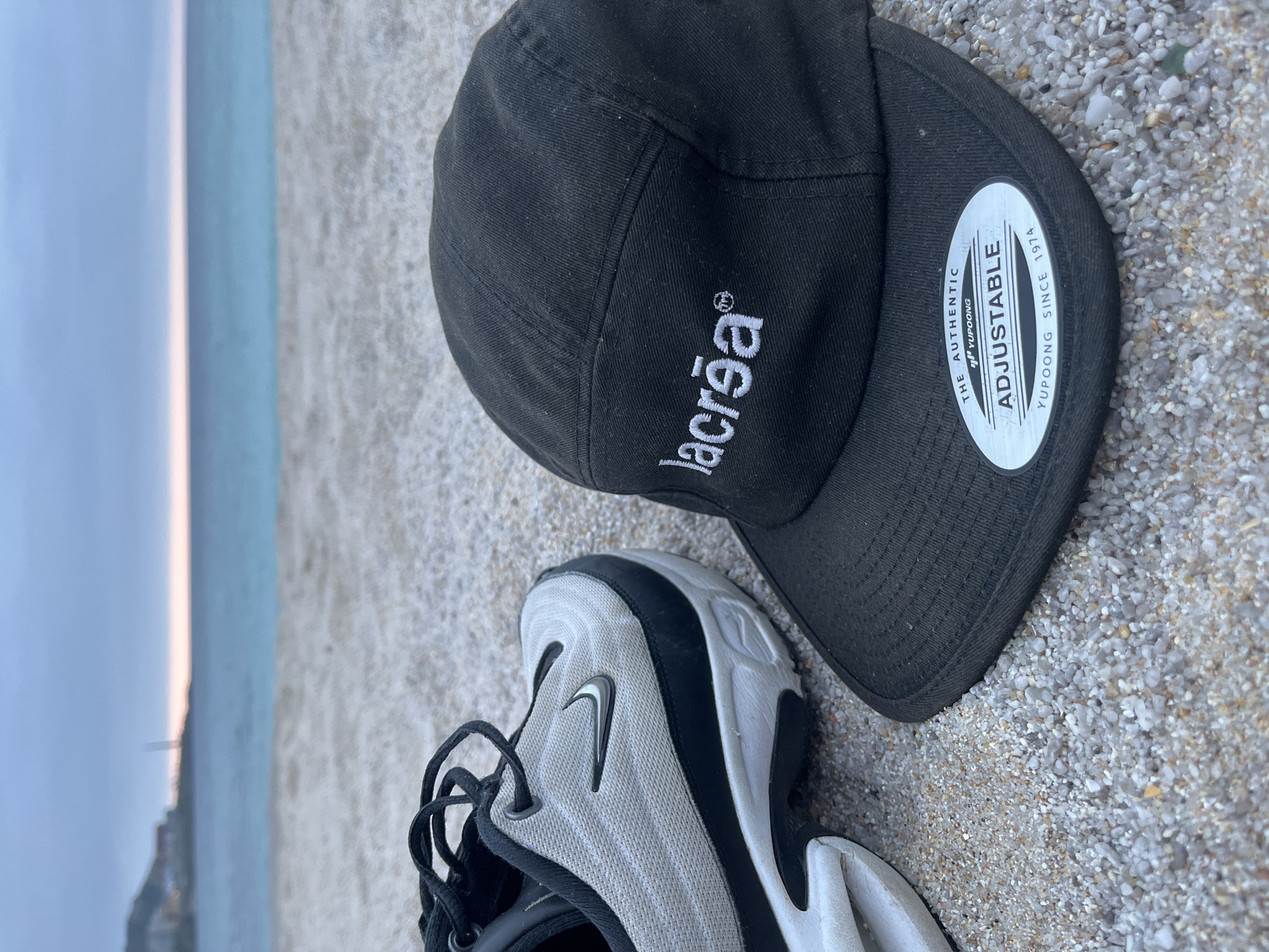 The image shows a black "lacreia" brand cap and a white and black Nike sneaker on a beach. The beach appears to be a small, pebble beach. In the background, one can see the sky and some of the city of A Coruña, Spain.