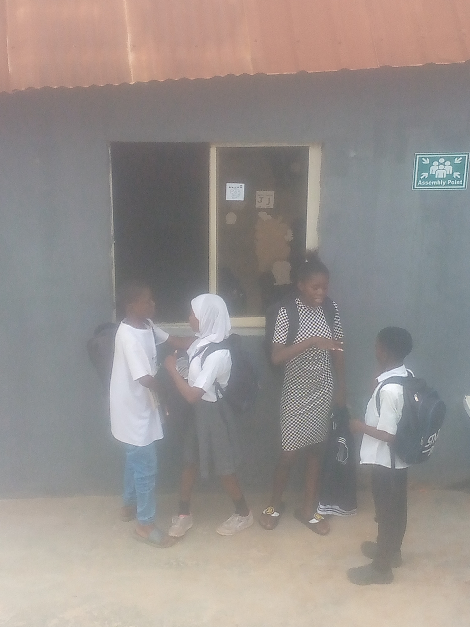 Four people are standing in front of a grey building with a brown roof. Two of the people on the left are wearing white shirts. One of them is wearing blue jeans and the other is wearing a grey skirt. The person in the middle is wearing a black and white dress. The person on the right is wearing black pants and a white shirt. All the people except the one in the black and white dress are wearing backpacks. There is a window behind the people and a green sign that says "Assembly Point" on the right side of the building. Location in the city of Unknown, Unknown.