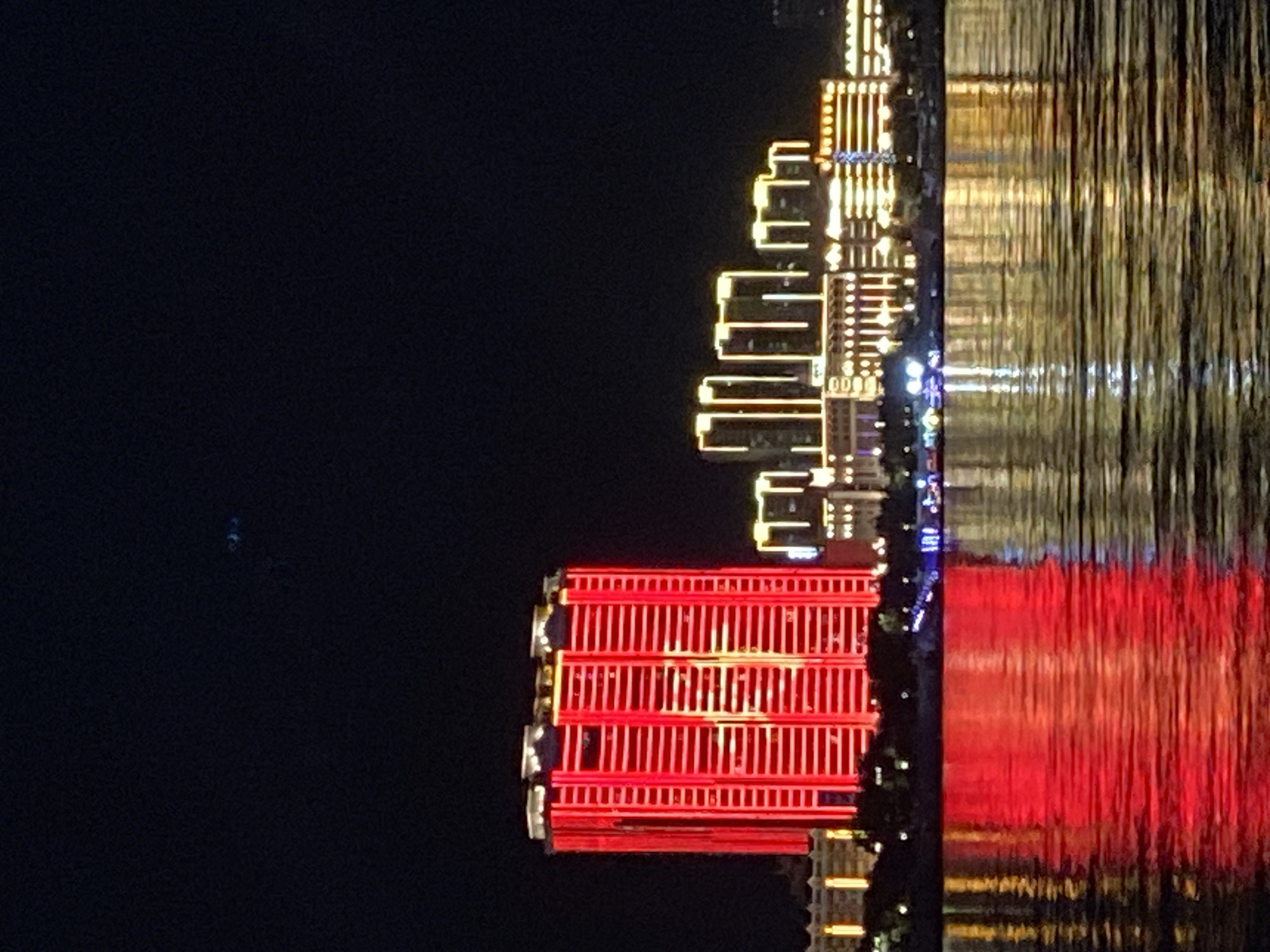The image shows a nighttime view of a city skyline across a body of water. The most prominent structure is a tall, rectangular building illuminated with bright red vertical lights. To the right of this building, a series of shorter, rectangular buildings are lit with yellow and white lights, outlining their structures. The buildings are situated along a waterfront, and their lights are reflected in the rippling water in the foreground. The sky above is dark, with a faint, blurry blue object visible in the upper left quadrant. The scene is likely of the Amur River embankment in Blagoveshchensk, Russia, which separates the city from China. The absence of identifiable individuals or specific actions suggests a scenic, urban landscape at night.