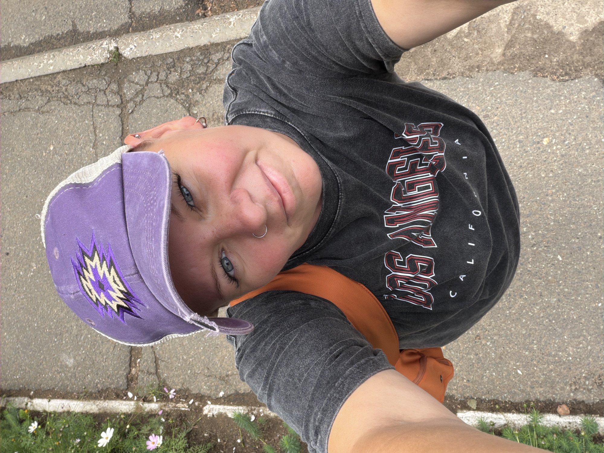 This is a selfie taken from a high angle, looking down at a person wearing a purple baseball cap with an embroidered emblem, a grey t-shirt with "LOS ANGELES CALIFORNIA" printed on it, and an orange strap visible. The person has blue eyes, a nose ring, and ear piercings. They are smiling at the camera. The setting appears to be outdoors on a paved surface with some greenery and small flowers along the edge. The sunlight suggests it is daytime. The image is rotated 90 degrees counter-clockwise.
