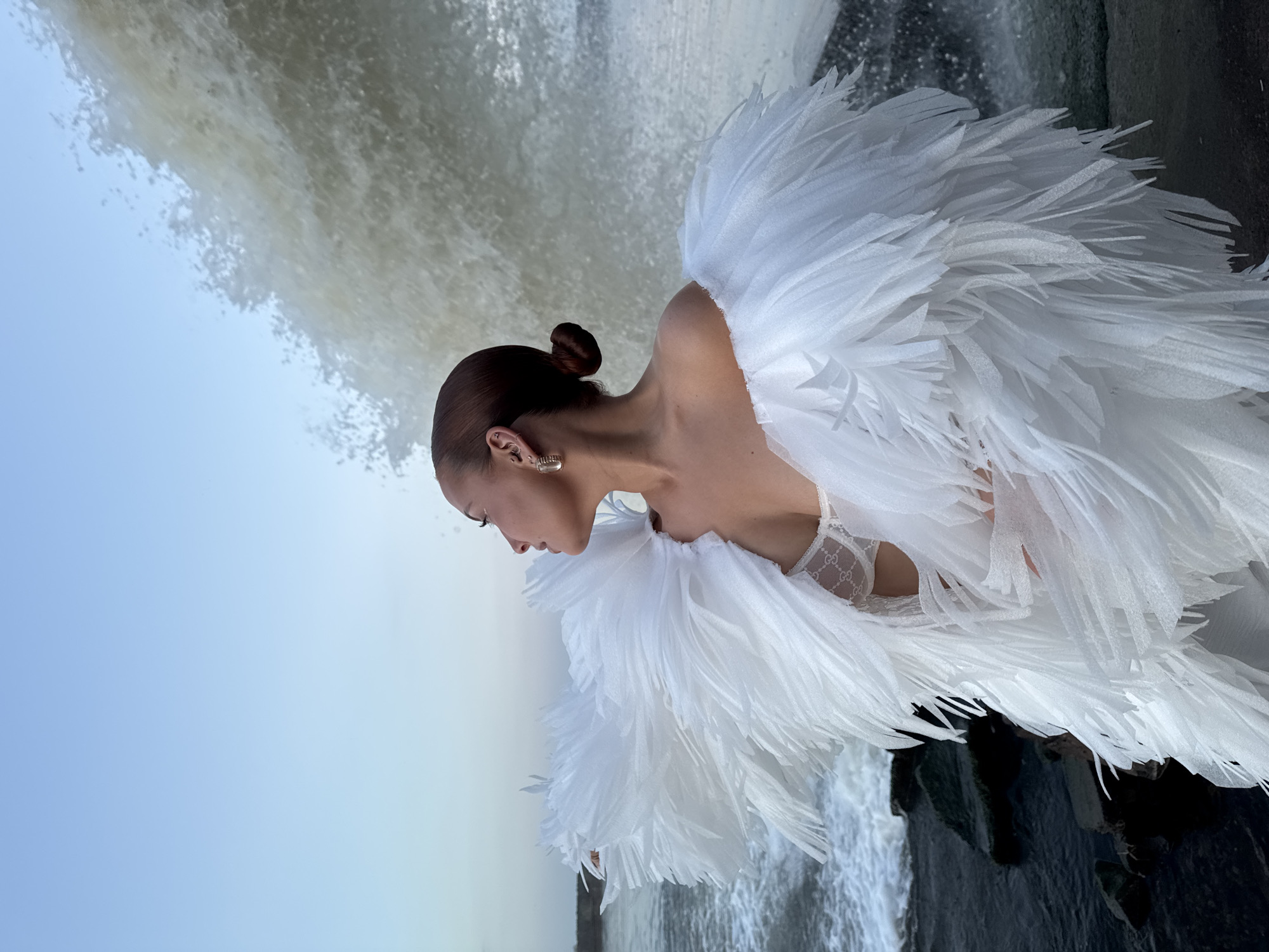 A woman with her hair in a bun is wearing a dress or garment with large, feather-like white embellishments around her shoulders. She stands in front of a waterfall. The setting is the city of Chaikovsky, Russia.